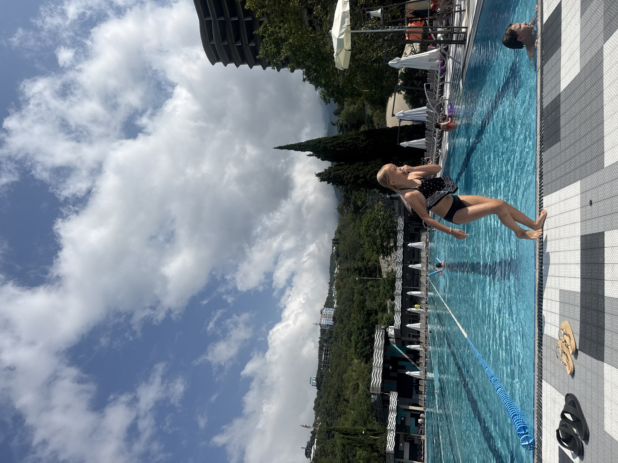 This image captures a scene at an outdoor swimming pool, likely in Yalta, Ukraine. The time of day appears to be daytime, with a partly cloudy sky.
The primary subjects are people engaged in activities around and in the pool. A young woman in a black and white patterned bikini is positioned in the foreground, seemingly mid-movement or posing at the edge of the pool. Further into the pool, another person, a young man, is visible in the water, his head and shoulders above the surface. In the distance, other individuals can be seen enjoying the pool.
The setting is a resort-like environment with a large, rectangular swimming pool. The pool is filled with clear blue water and marked with lanes by a blue rope. The pool deck is paved with a patterned material, and lounge chairs and white umbrellas are arranged along the side, suggesting a place for relaxation. Beyond the pool, lush green trees and hills form the backdrop, with a prominent, tall, dark green coniferous tree standing out. A large, circular, unfinished building structure is visible in the upper left, adding a unique architectural element to the landscape.
Notable details include the clear blue water of the pool, the textured pattern of the pool deck, and the scattered clouds against a blue sky. The overall atmosphere seems relaxed and summery. There is no visible text in the image.