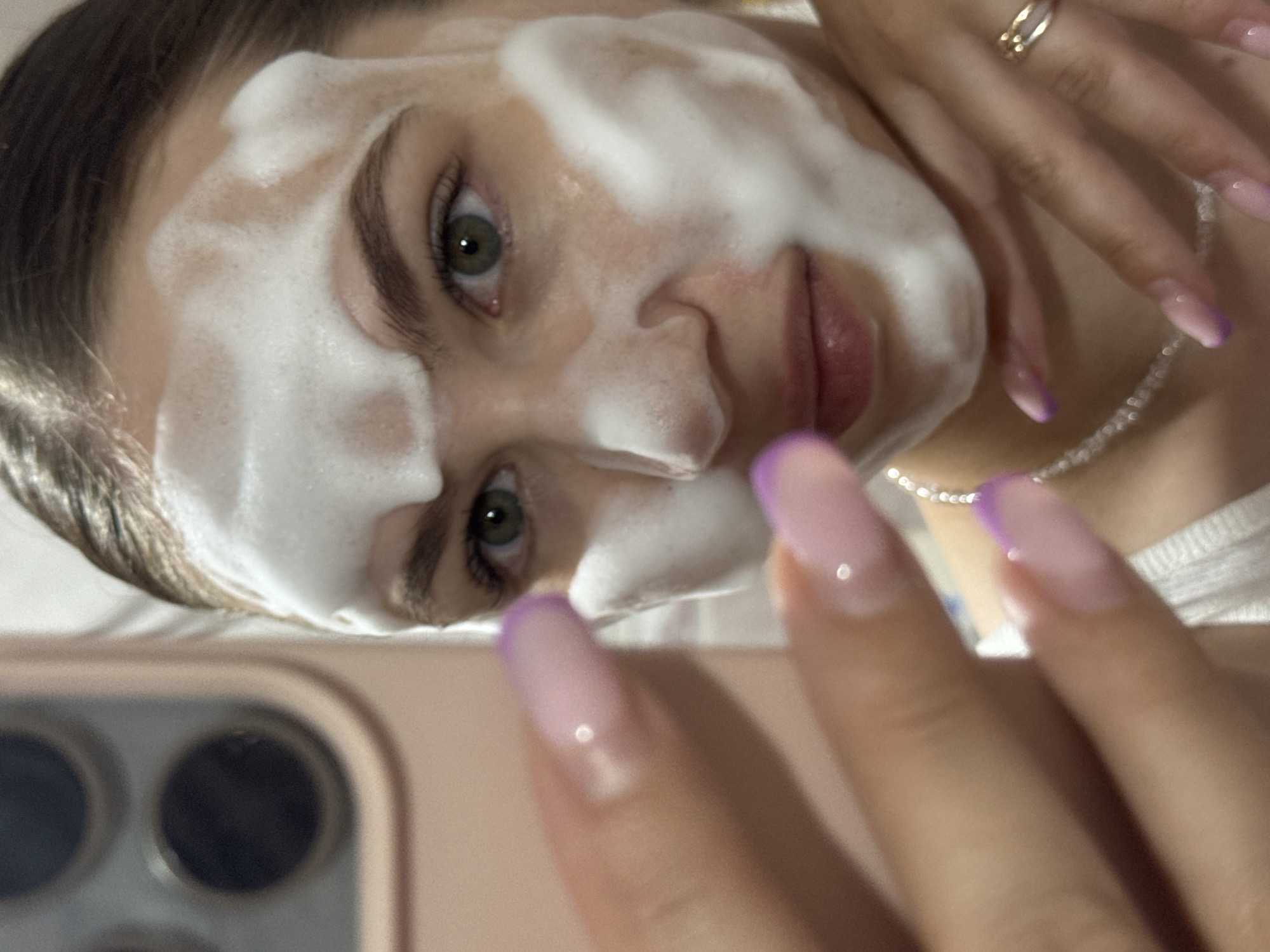 A young woman with green eyes and dark hair, her face covered in white foam, is captured in a close-up shot. She is wearing a white top and a pearl necklace. Her hands, with long, light purple French manicured nails, are visible in the foreground and near her neck. One hand is held up, obscuring part of her face, and the other is resting on her chest, adorned with a ring on her right ring finger. The background is blurred, showing what appears to be the back of a pink phone with multiple camera lenses on the left side. The lighting is soft, highlighting the foam and her facial features. The image does not provide any information about the location in the city of Chaikovsky, Russia.