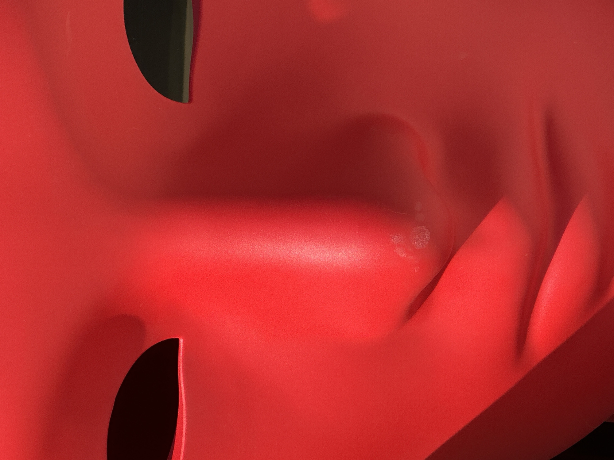 A close-up photograph shows a section of a red, possibly plastic, object with a curved and textured surface. There are two dark, irregular openings visible, suggesting the object is a mask or similar form. The lighting is uneven, creating highlights and shadows across the surface. Minor surface imperfections are visible. The location and specific details about Budva, Montenegro are not discernible from the image.