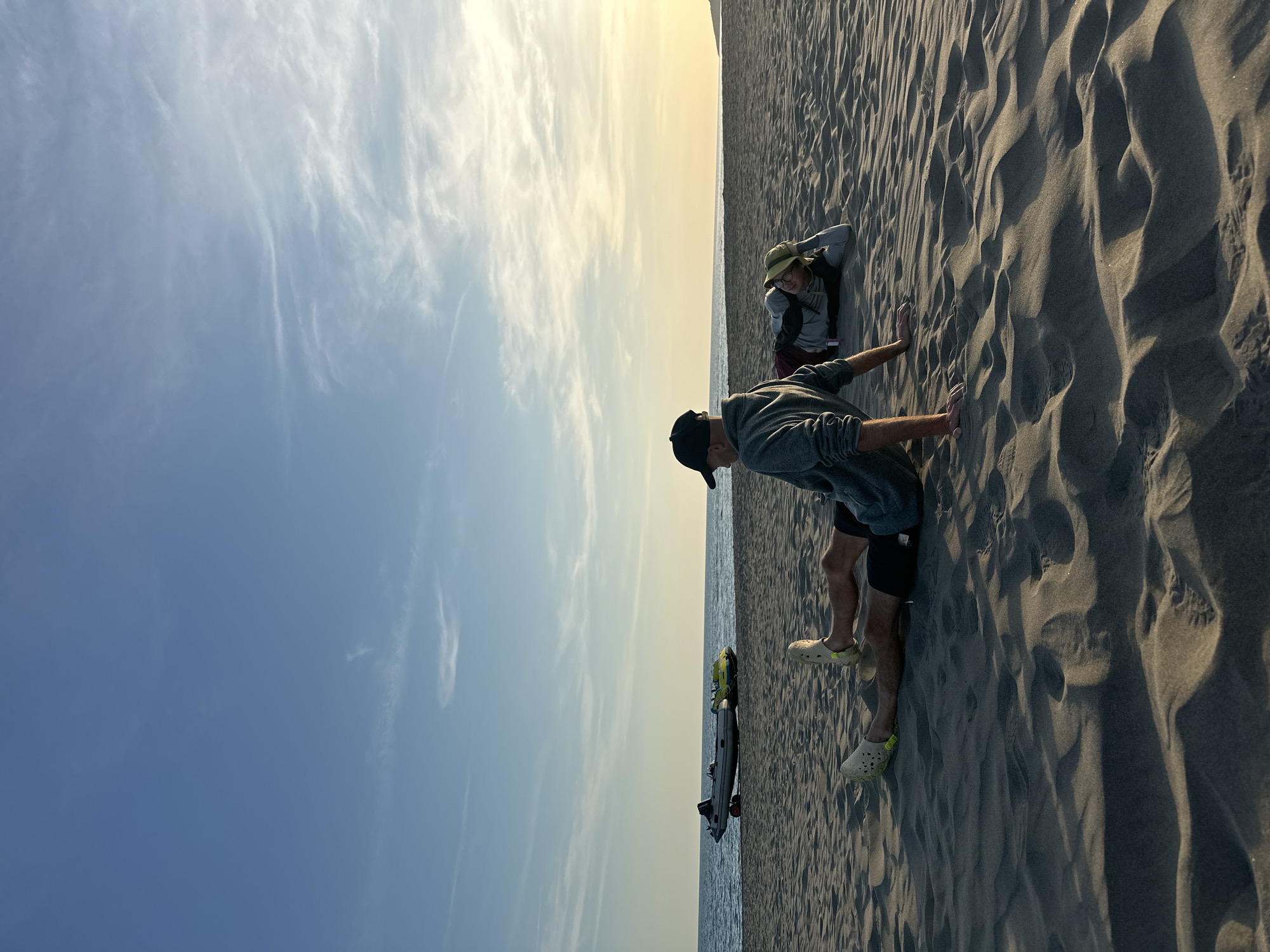 The image captures a scene on Velika Plaža (Long Beach) in Donji Štoj, Montenegro. The sky transitions from a pale blue to a faint yellow near the horizon, marked by wispy clouds. Below the sky, a strip of water is visible, indicating the Adriatic Sea.
On the sandy beach, two people are present. A man wearing a dark baseball cap, a gray sweatshirt, black shorts, and light-colored Crocs with yellow accents stands with his hands on the sand, leaning forward. Behind him, a person wearing a hat and a long-sleeved shirt is partially obscured in the sand. A sandboard with visible yellow and gray elements sits near the edge of the water. The sand itself is heavily textured, with prominent wave-like formations and visible footprints.