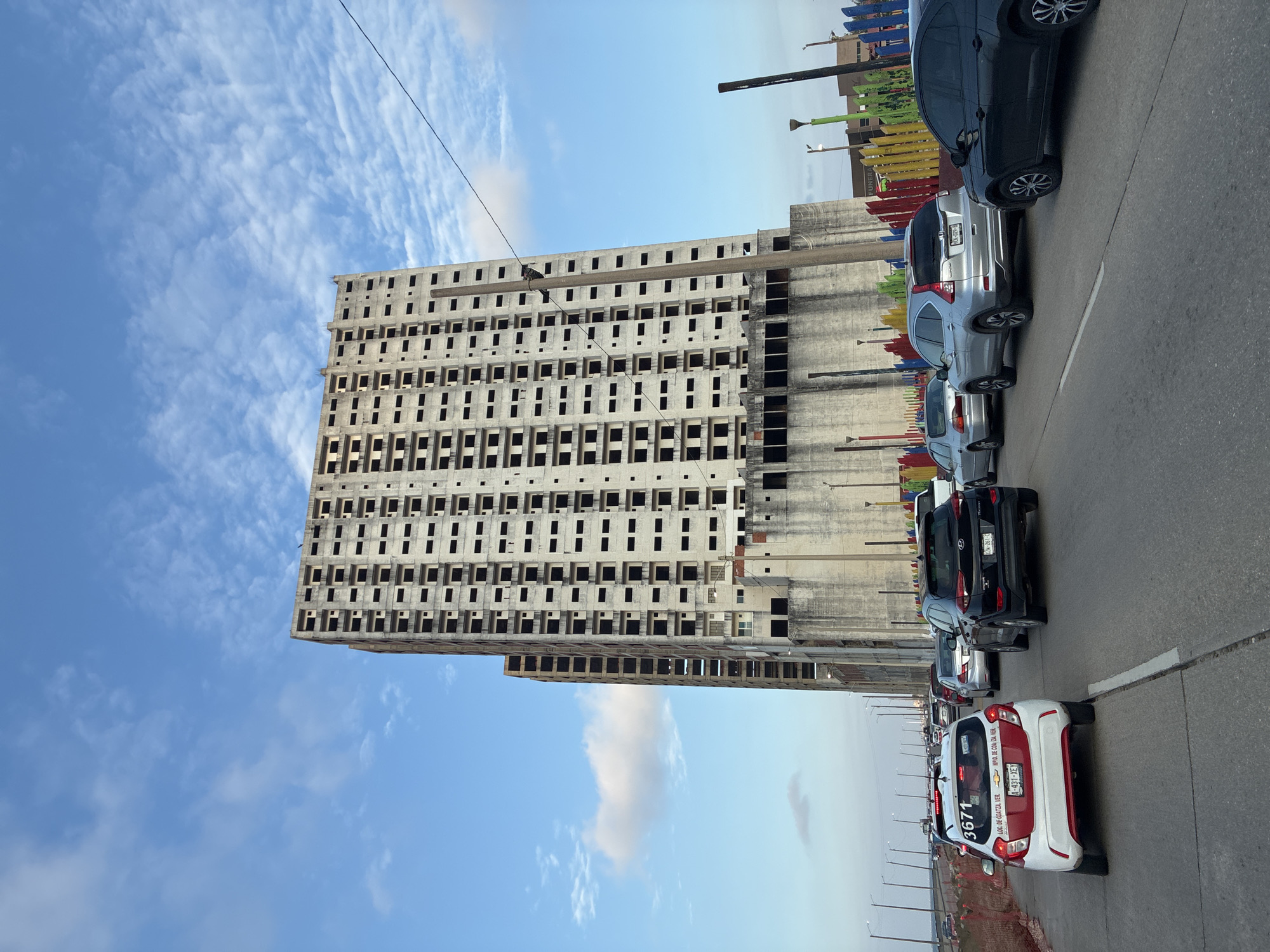 A vertical, wide shot showcases an unfinished concrete building against a blue, partially cloudy sky. The building features multiple rows of empty window spaces, a stark contrast to its gray, unadorned facade. To the right, cars are parked along a gray asphalt road. A red-and-white taxi with the license plate "A-431-XEY", bearing the number "3671" and the text "LOC DE COATZA VER" is in the foreground. The license plate "XZW-261-C" is visible on the black car behind. The location is Coatzacoalcos, Mexico.