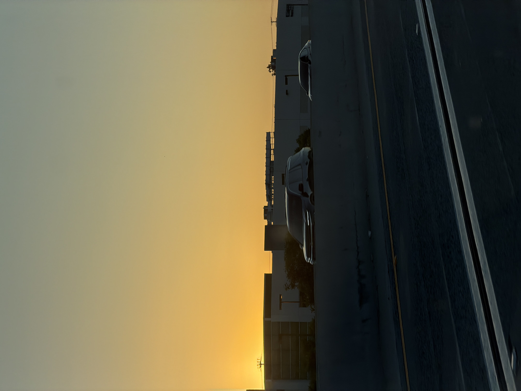 This image depicts a sunset over the city of Burbank, United States of America. The sky transitions from a bright orange near the horizon to a muted yellow and then a pale green higher up. Silhouetted against the sky are buildings that appear to be warehouses or industrial structures. In the foreground, a dark, shadowy barrier runs horizontally across the lower half of the frame, suggesting the viewer is on an elevated roadway, possibly a freeway. Two vehicles are visible in the middle ground, driving on a road behind the barrier. The vehicle on the left is a dark-colored SUV, while the vehicle on the right is a darker sedan. The road surface in the immediate foreground is dark and appears to be asphalt, with a faint yellow line marking a lane divider. The overall impression is a common urban landscape at dusk.