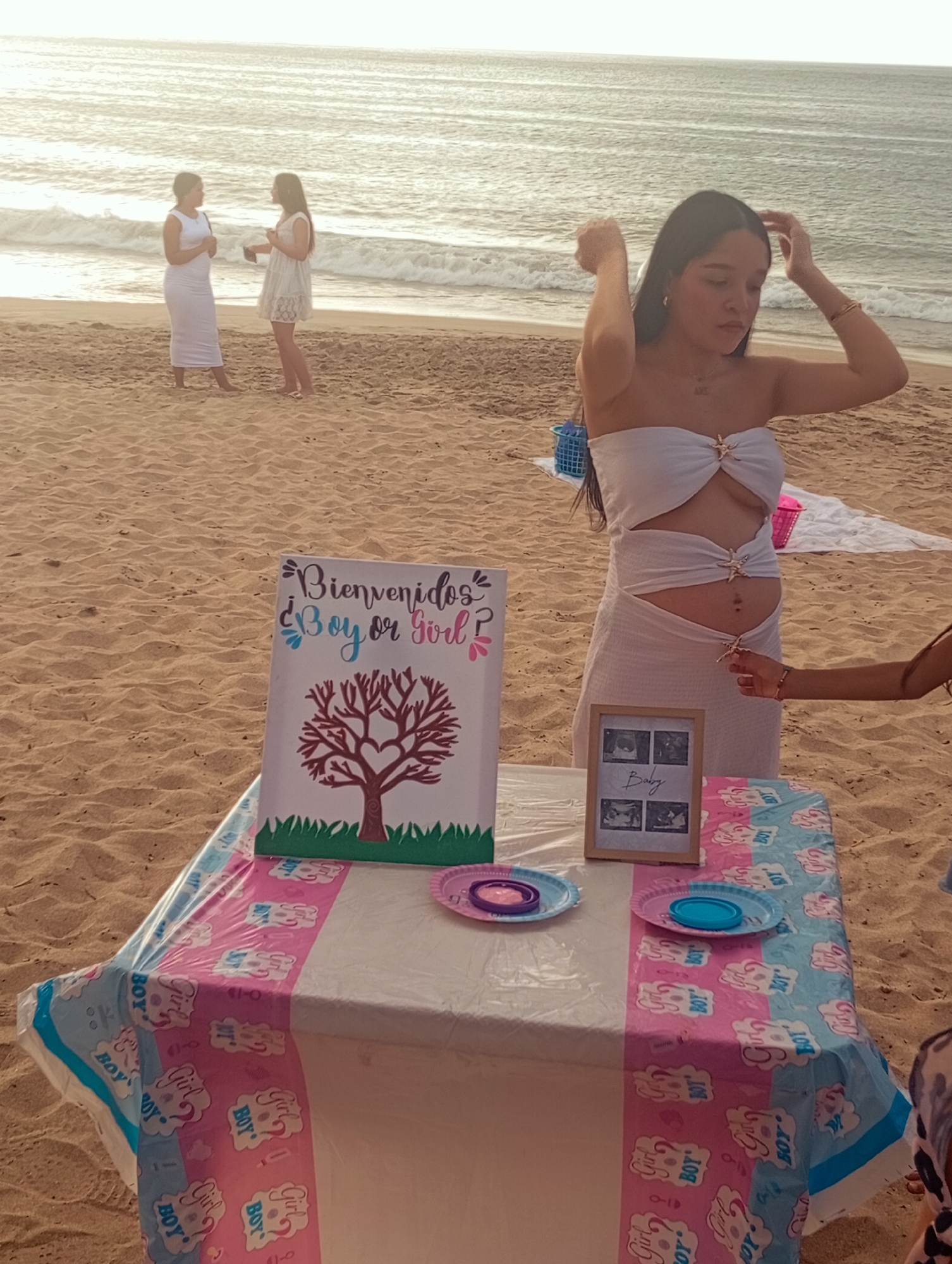 The image depicts a beach scene, likely a gender reveal celebration. A pregnant woman in a white outfit stands near a table covered with a tablecloth printed with "Boy or Girl" motifs in pink and blue. The table holds a sign that reads "Bienvenidos ¿Boy or Girl?" in Spanish and features a tree graphic. Additionally, there are sonogram photos in a frame, plates, and a table cloth. In the background, two women in white dresses are seen conversing near the water's edge. The scene appears to be taking place on a beach in Pedro González, Venezuela, considering the location was specified. The sky is bright, suggesting it is daytime, and the overall atmosphere is celebratory.