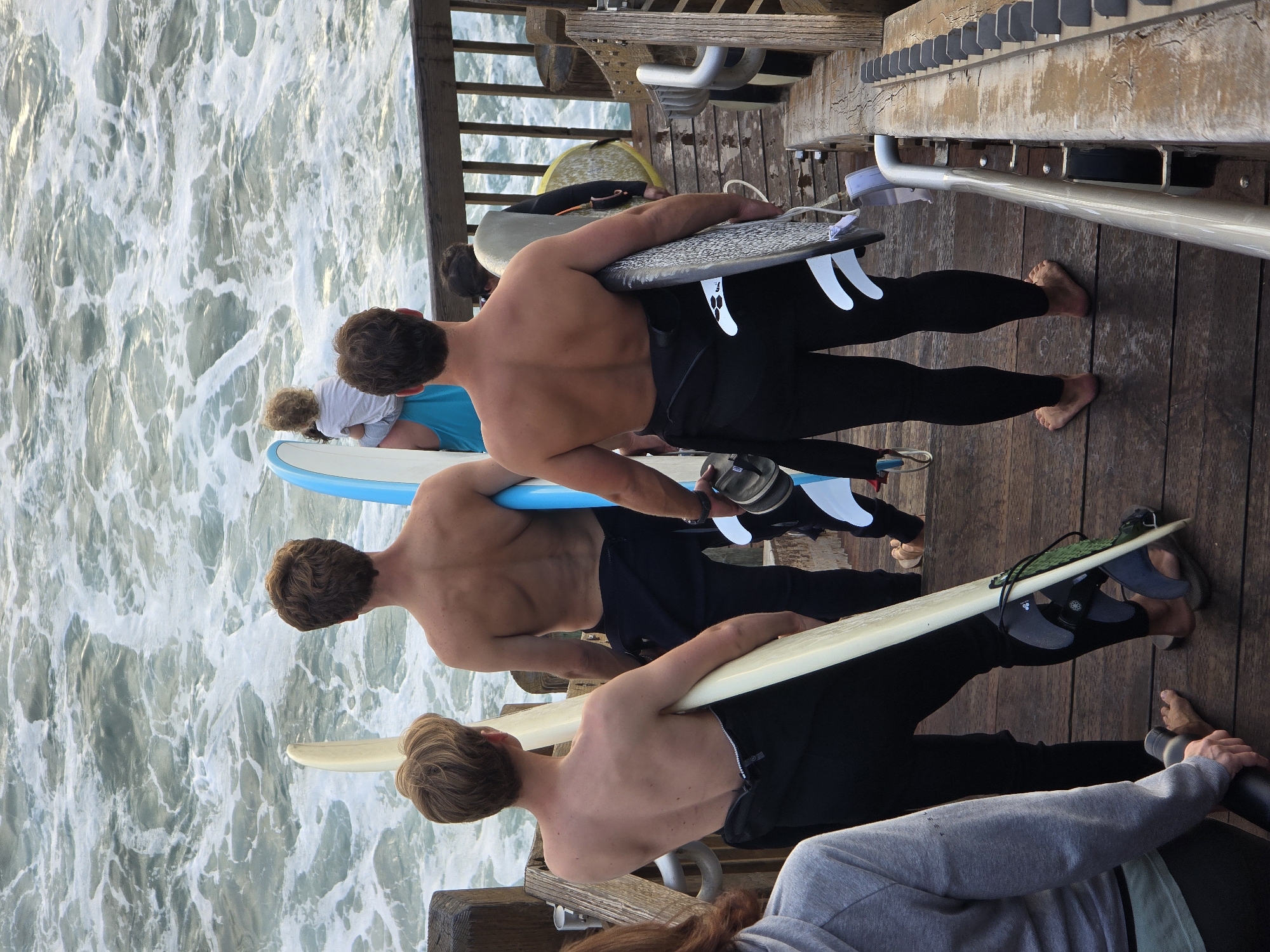 In Encinitas, California, a group of individuals, primarily surfers, are positioned on a wooden boardwalk adjacent to the ocean. The ocean is rough with significant white water. Several people are holding surfboards and appear to be preparing to enter or having just exited the water. One person in the foreground is wearing a grey hoodie and black pants, holding a black water bottle. To their right, a surfer in a black wetsuit carries a light-colored surfboard. Further back, another surfer, shirtless, stands with a blue surfboard. A child is being held by an adult in the background, also near a surfboard. The boardwalk has wooden planks and a metal railing. The scene suggests a surfing activity is either underway or about to commence.