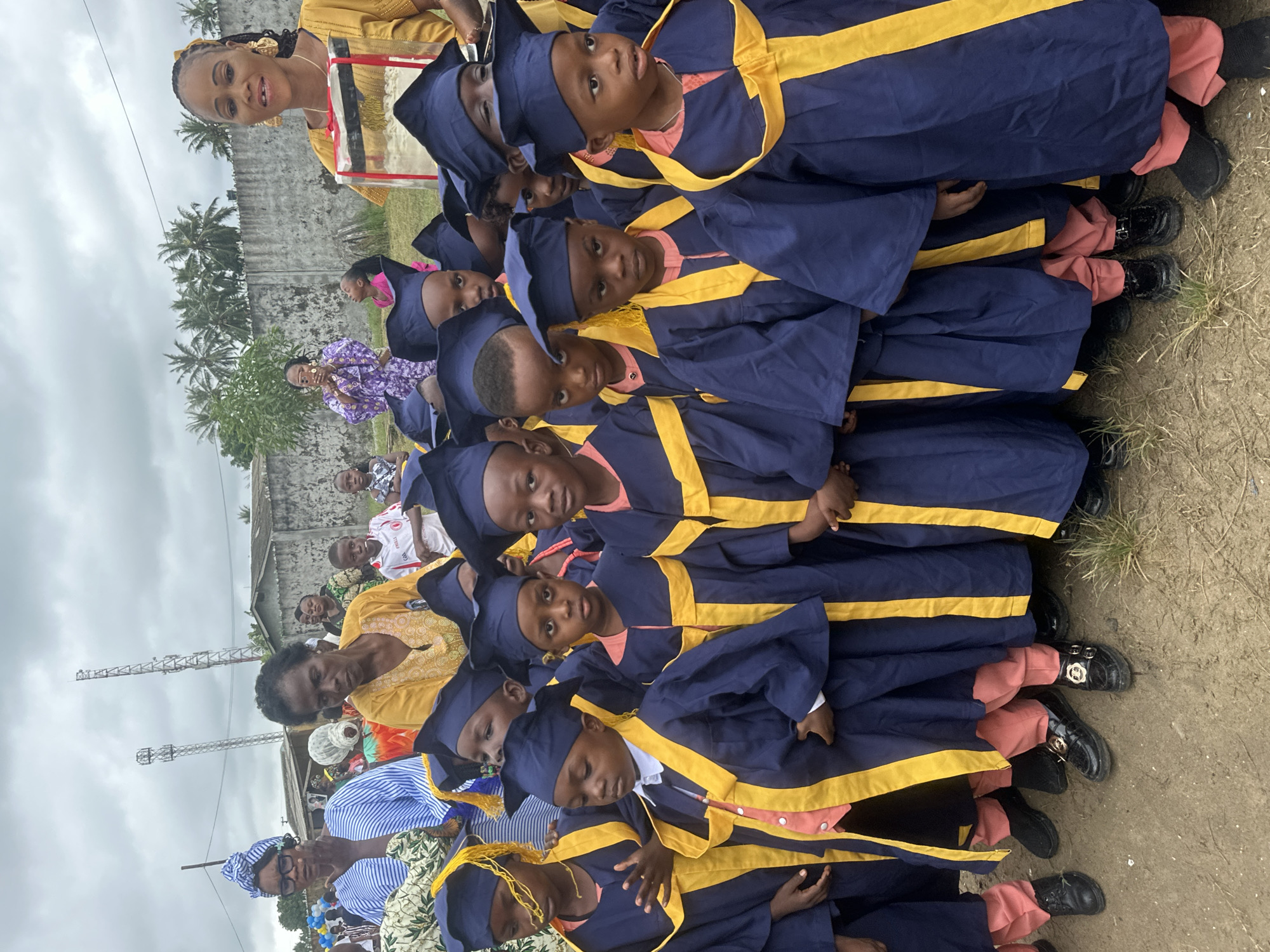 A group of young children in blue graduation gowns and caps with yellow trim stand in rows, facing forward. Many of them are looking at the camera. In the background, several adult women are present, some wearing colorful clothing. One woman in a yellow dress holds a box wrapped in clear plastic. Behind the people, there is a grey concrete wall, palm trees, and some structures that appear to be communication towers. The ground is dirt. The scene is outdoors, under an overcast sky. The image appears to have been taken in Badagry, Nigeria, likely at a graduation ceremony.