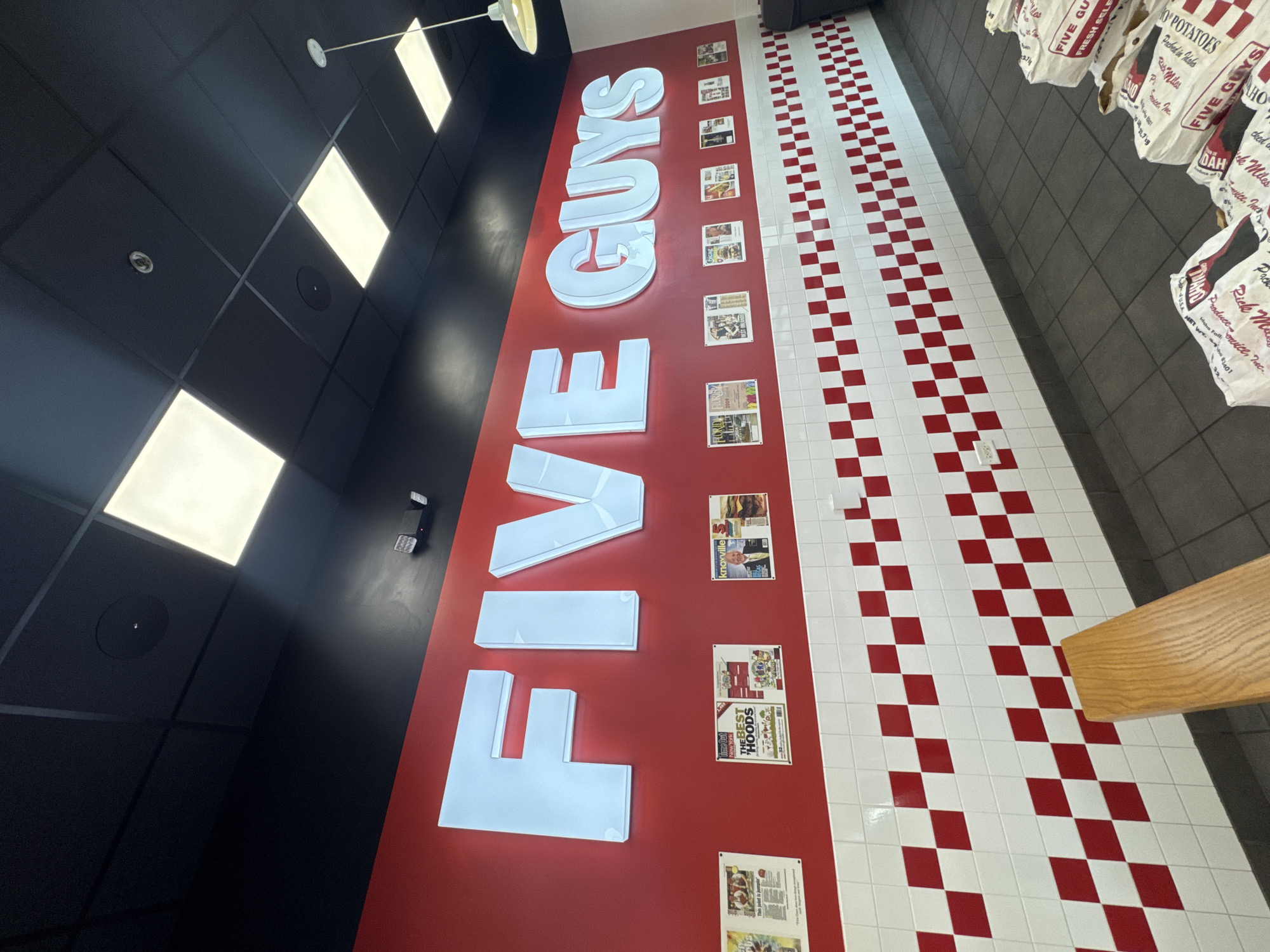 This image shows the interior of a Five Guys restaurant. The restaurant's logo, "FIVE GUYS," is prominently displayed in large, illuminated white letters on a red wall. Newspaper clippings and magazine articles are pinned to the wall beside the logo. The floor has a red and white checkered pattern. To the right of the floor is a gray tiled wall with brown bags hanging on it. The black ceiling above has square, recessed lights. There is no indication of its location in Bloomingdale, United States of America.