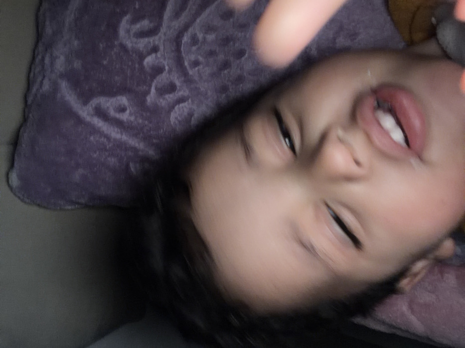 This image depicts a young child, possibly a toddler or preschooler, captured in an indoor setting in Dar Bouazza, Morocco. The child, with dark hair, light-to-medium skin, and an unclear gender, is positioned centrally in the frame. Their eyes are partially closed or squinting, and their mouth is slightly open, revealing several upper and lower teeth. A whitish substance is visible around the lips, suggesting food or drink residue. A blurred finger, likely belonging to the child or another individual, is present in the foreground on the right, partially obscuring the child's face. The child appears to be lying down on a soft surface, possibly a bed or sofa. The background consists of a dark purple, textured blanket or pillow with an indistinct pattern, visible to the upper right and behind the child's head. A darker, unidentifiable area is visible to the upper left. The lighting is dim, and the overall image exhibits slight motion blur, particularly in the background and foreground finger.