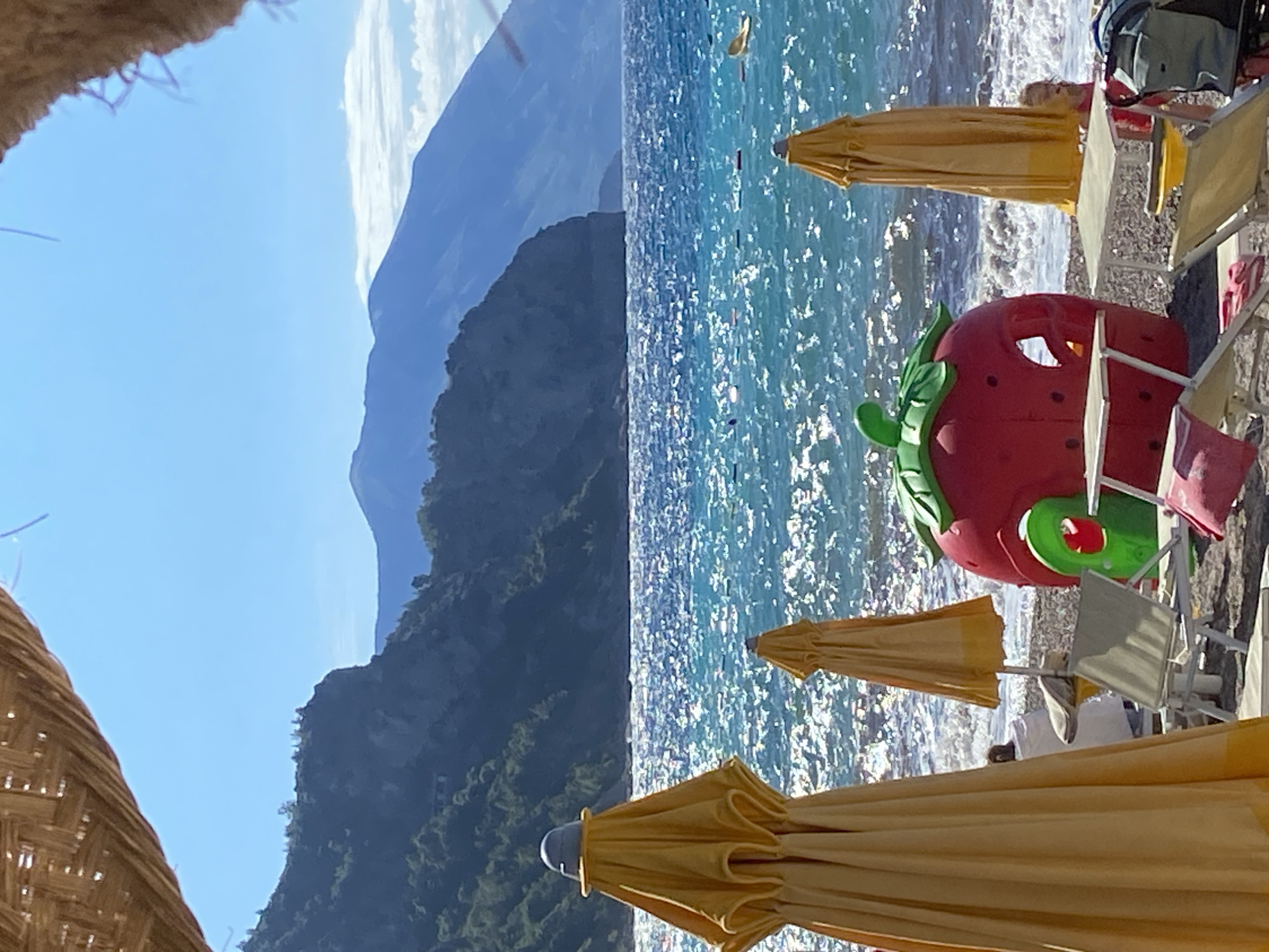 A low-angle, medium shot captures a sun-drenched beach scene in Prijevor, Montenegro. The foreground is dominated by the tops of two yellow beach umbrellas, one of which is partially obscuring a child in a red swimsuit in the right background. In the center of the frame, a large, red, strawberry-shaped playhouse is situated on the pebbled beach, with a green slide protruding from its side. Several empty white lounge chairs are arranged around the playhouse. Beyond the beach, the sparkling turquoise sea stretches out, with faint outlines of people swimming and a yellow float visible. The horizon is defined by a series of majestic, blue-tinged mountains, their slopes covered in lush green vegetation. The sky above is a clear, brilliant blue, with a woven canopy framing the top of the image.