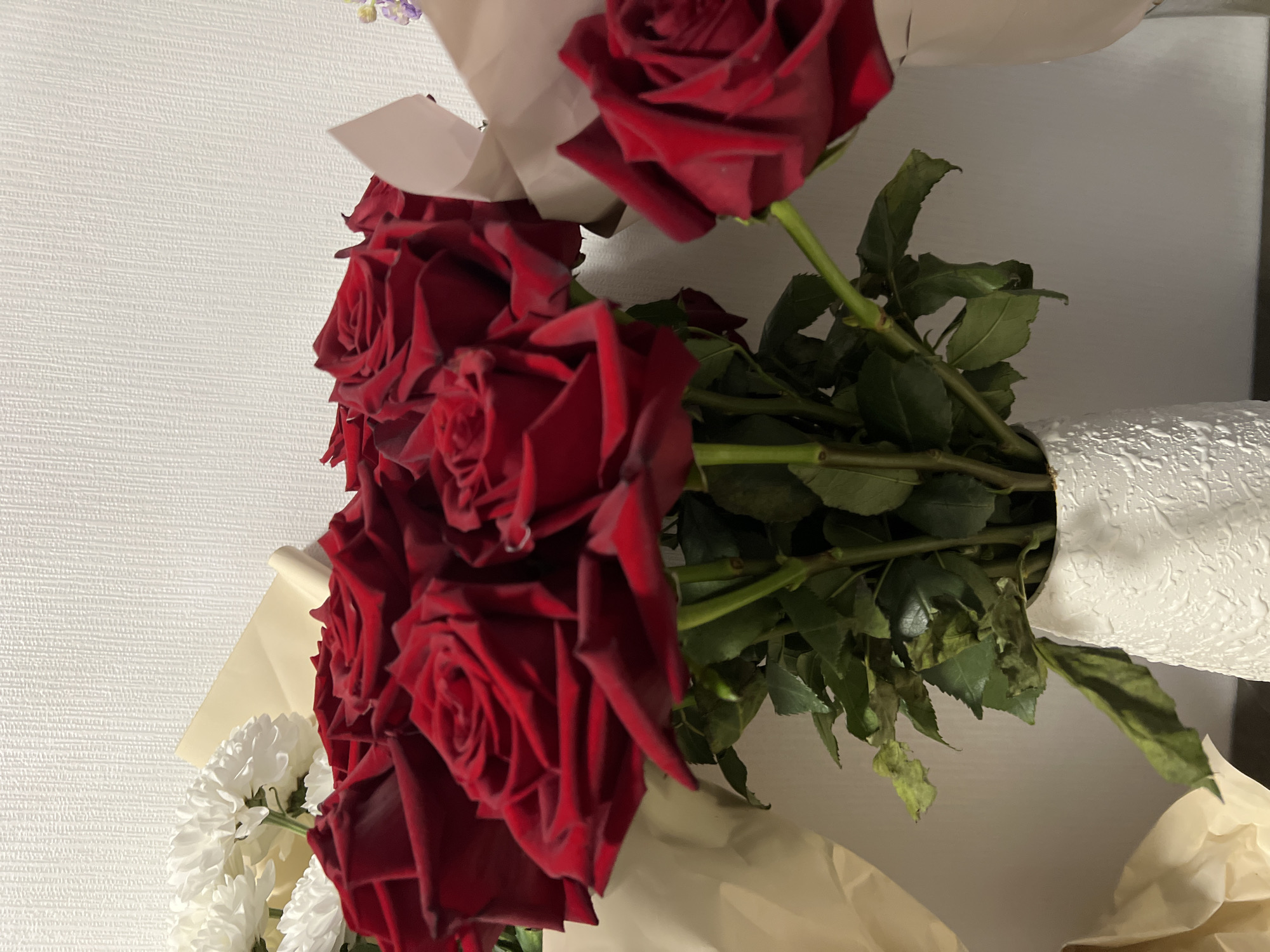 A bouquet of red roses is displayed in a textured white vase. The bouquet is partially wrapped in light brown paper. To the left of the red roses, a bouquet of white chrysanthemums is visible. The background is a textured, off-white wall. There are no people present in the image, and no specific location in Yekaterinburg, Russia can be identified. The image is a still life of flowers.