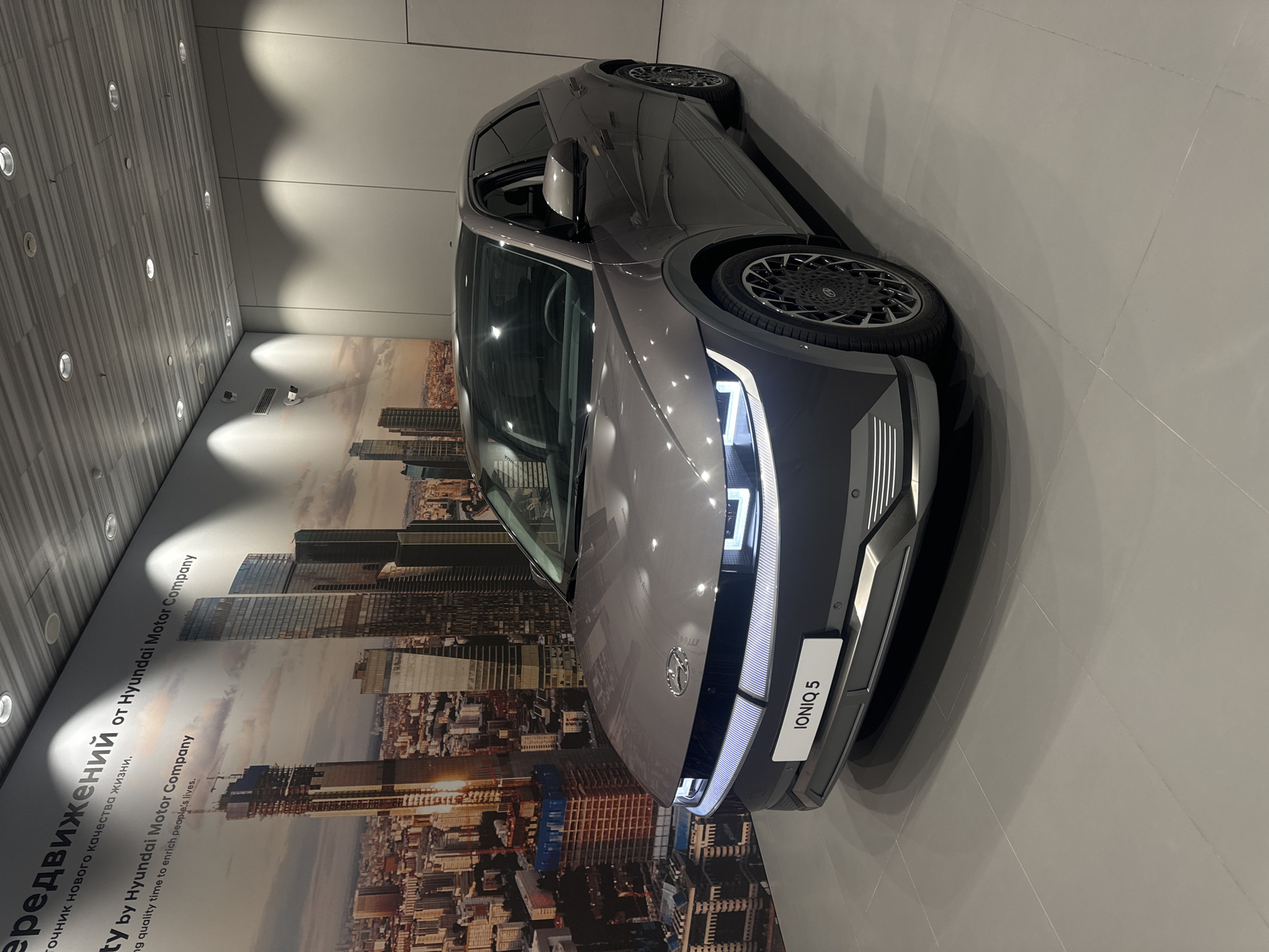 Here is a description of the image:
The image shows a gray Hyundai Ioniq 5 car parked indoors. The car is positioned in front of a large mural depicting a cityscape, identified as Moscow, Russia, based on the architectural style of the buildings. The mural also features text in both Russian and English, mentioning "Hyundai Motor Company." The flooring is a light gray tile. The overhead lighting suggests the car is in a showroom or a promotional space. There are no people visible in the image.