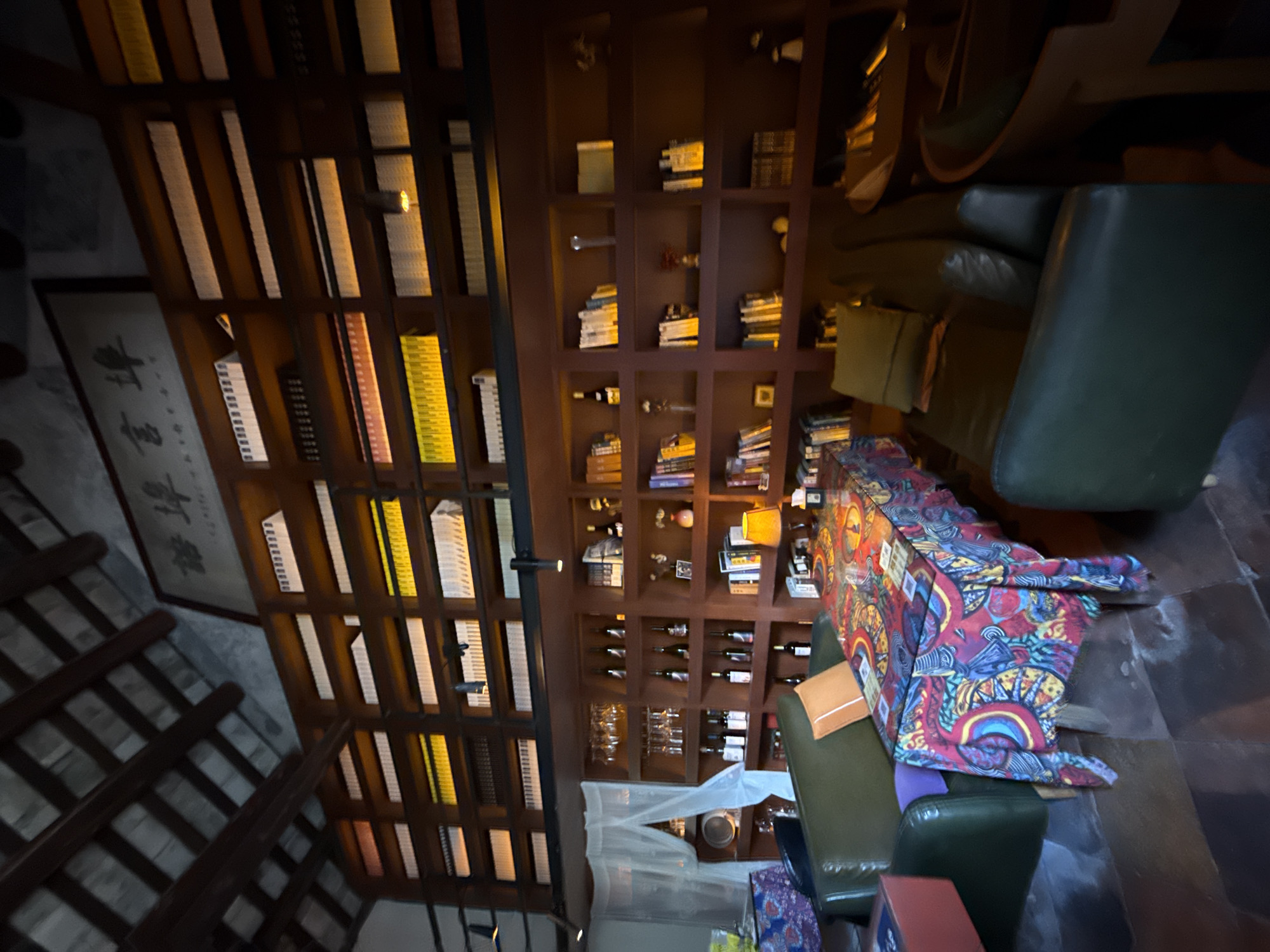 The image shows an interior shot of a room that appears to be a library or a lounge area within a building in Longgang District, China. The room features extensive wooden bookshelves filled with books, bottles, and decorative items, stretching from floor to ceiling. The ceiling has exposed wooden beams. In the foreground, there is a dark green leather sofa and armchair. A rectangular table is placed between them, covered with a vibrant, patterned tablecloth. The table also has some items on it, possibly drinks or menus. The lighting in the room is warm and ambient, with some shelves illuminated by soft lights, creating a cozy atmosphere. A white sheer curtain is visible on the left side of the image, near the sofa.