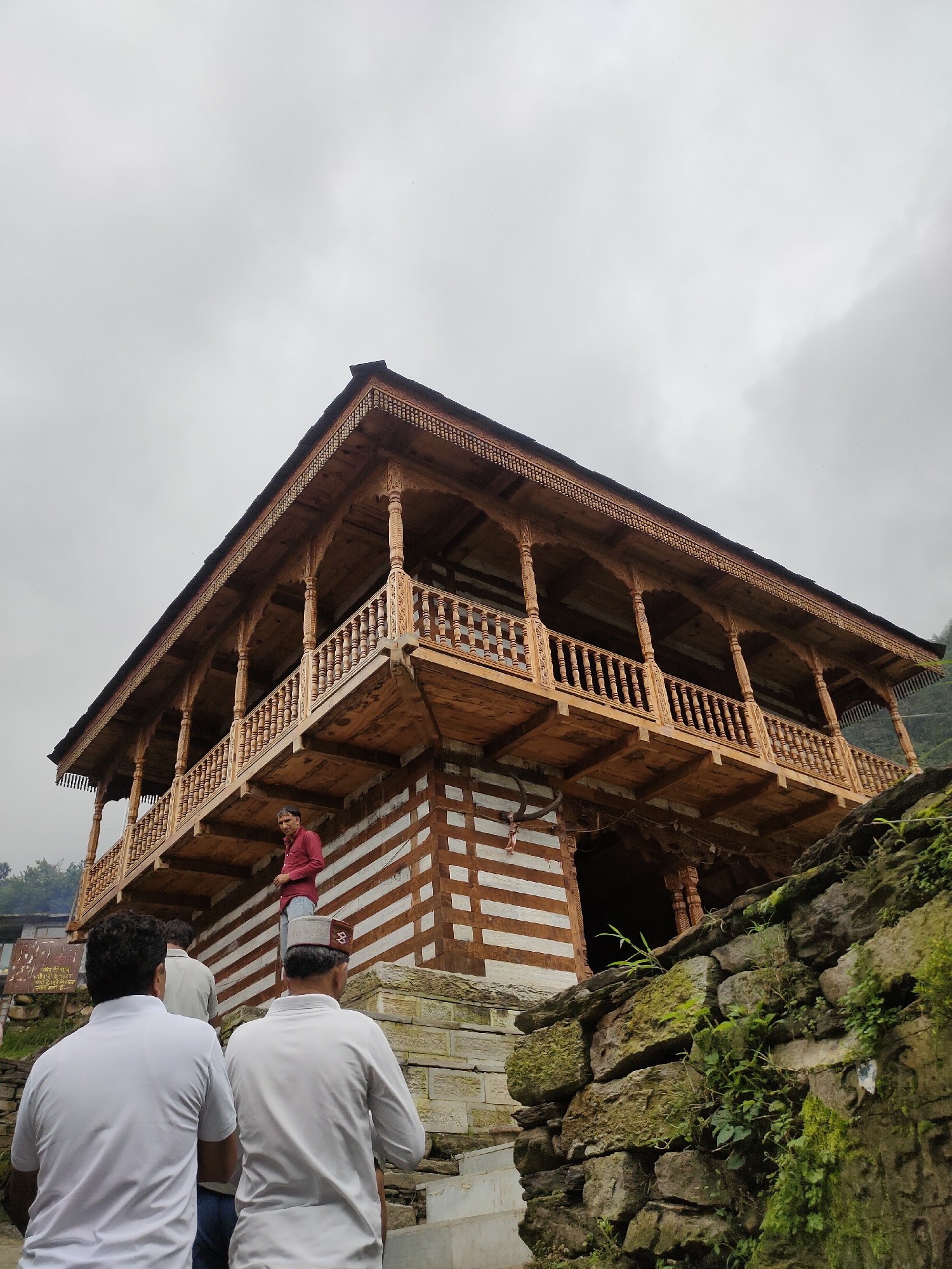 The image depicts a traditional wooden building in Damadi, India, with a cloudy sky overhead. In the foreground, three individuals are visible from behind, walking up a stone staircase. The building itself features intricate wooden carvings on its balconies and roofline, with a distinct pattern of white and brown stripes on its lower facade. A man in a red shirt and jeans stands on the upper balcony, looking towards the left. The surrounding environment includes a stone wall covered in moss and vegetation on the right side of the image, and glimpses of greenery in the background. A sign with text in Hindi is also partially visible on the left. The architectural style suggests a historical or cultural significance of the structure.