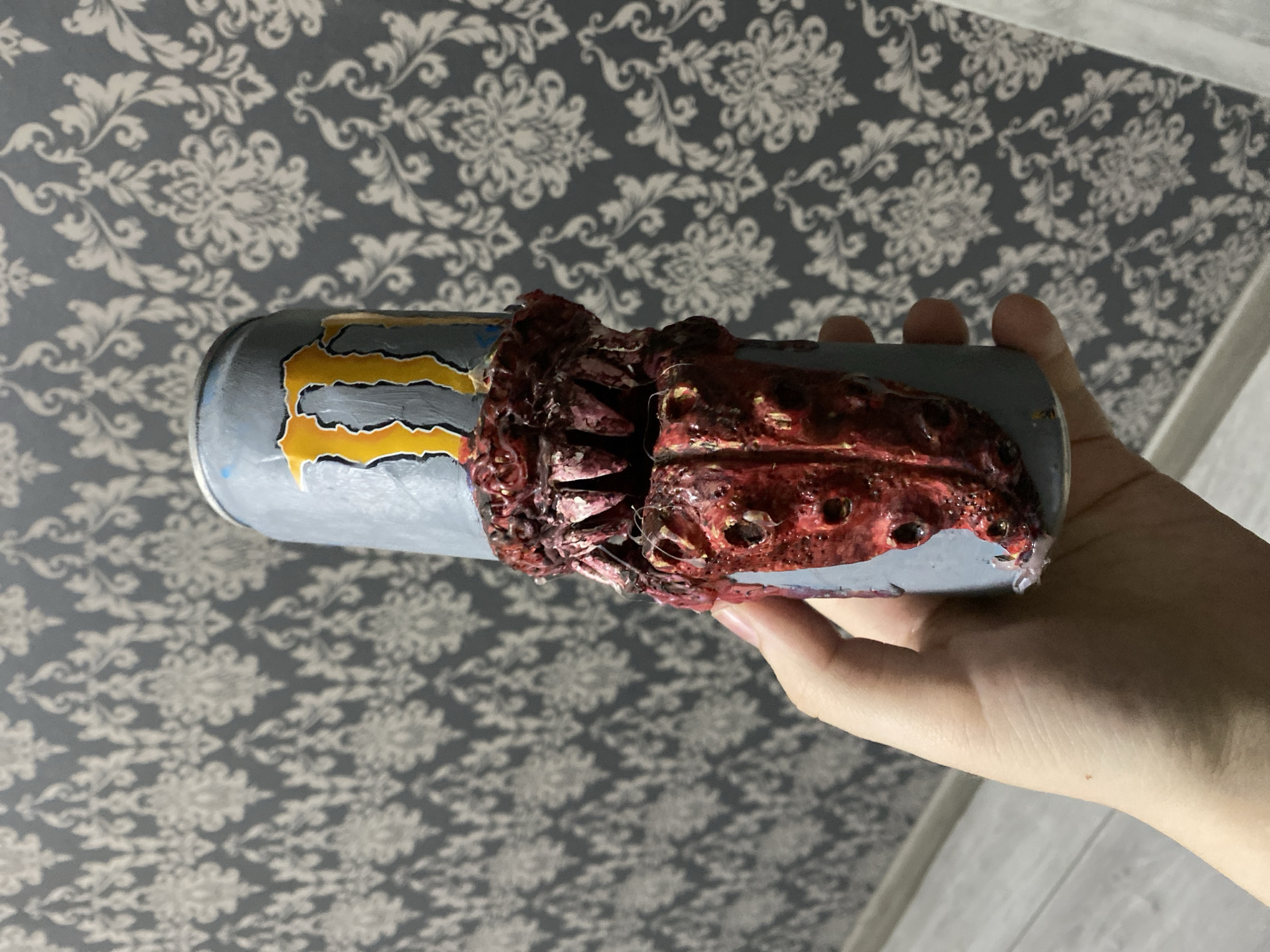A hand holds a modified Monster energy drink can. The can is painted grey and a stylized yellow "M" is visible. Protruding from the can is a red, textured material resembling fleshy tendrils or gore. The background is a grey patterned wallpaper. The image does not contain the specific location within the city of Ternopil, Ukraine.