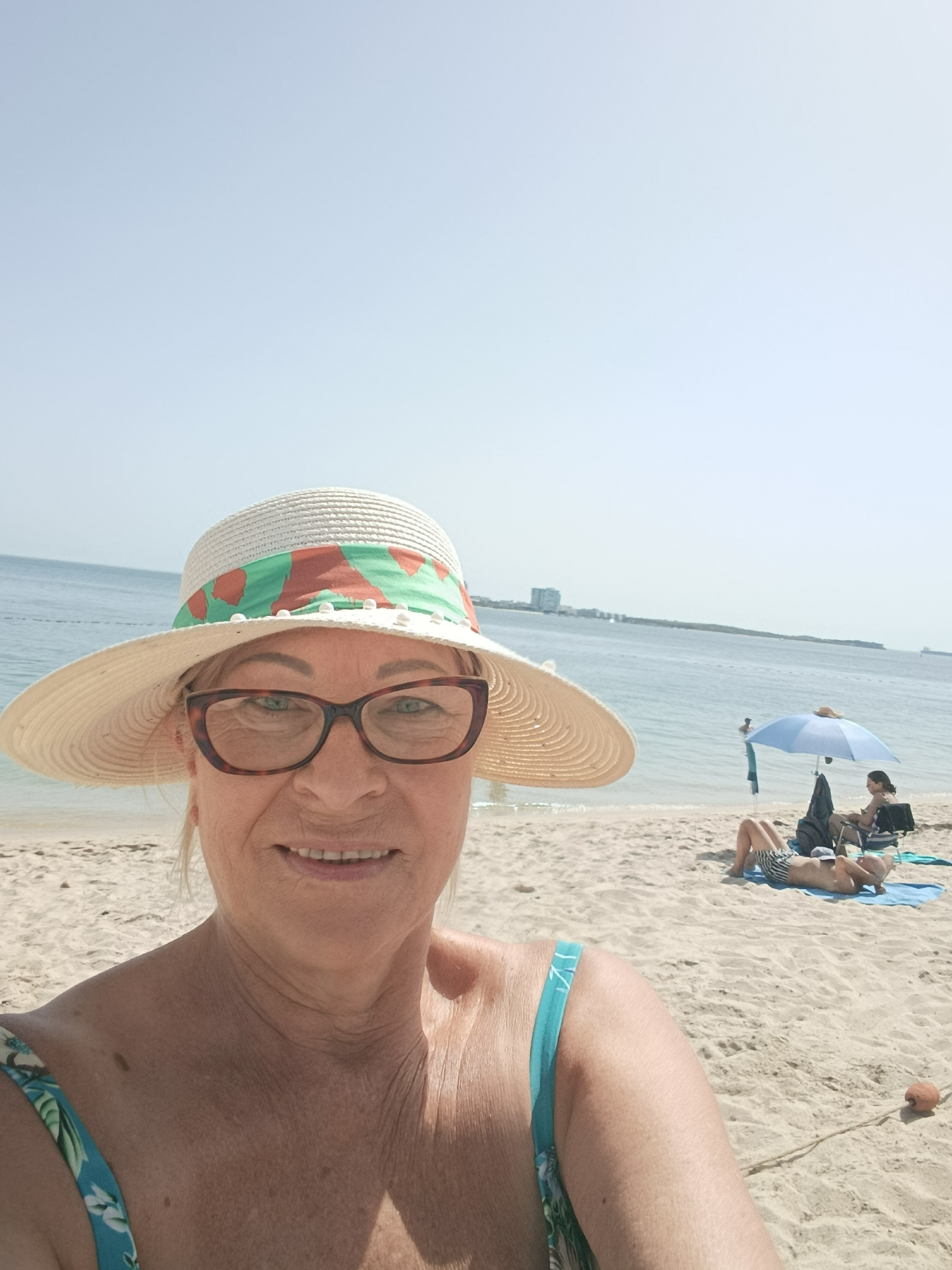 A woman, wearing a wide-brimmed hat and glasses, takes a selfie on a beach. She has blonde hair and is smiling at the camera. Behind her, the ocean stretches out to the horizon, with a faint cityscape visible in the distance. Two people are relaxing on the sand under a blue umbrella, and another person is swimming in the ocean. The beach is made of light-colored sand, and the water is calm. The sky is clear and bright. The overall impression is one of a relaxed and sunny day at the beach.