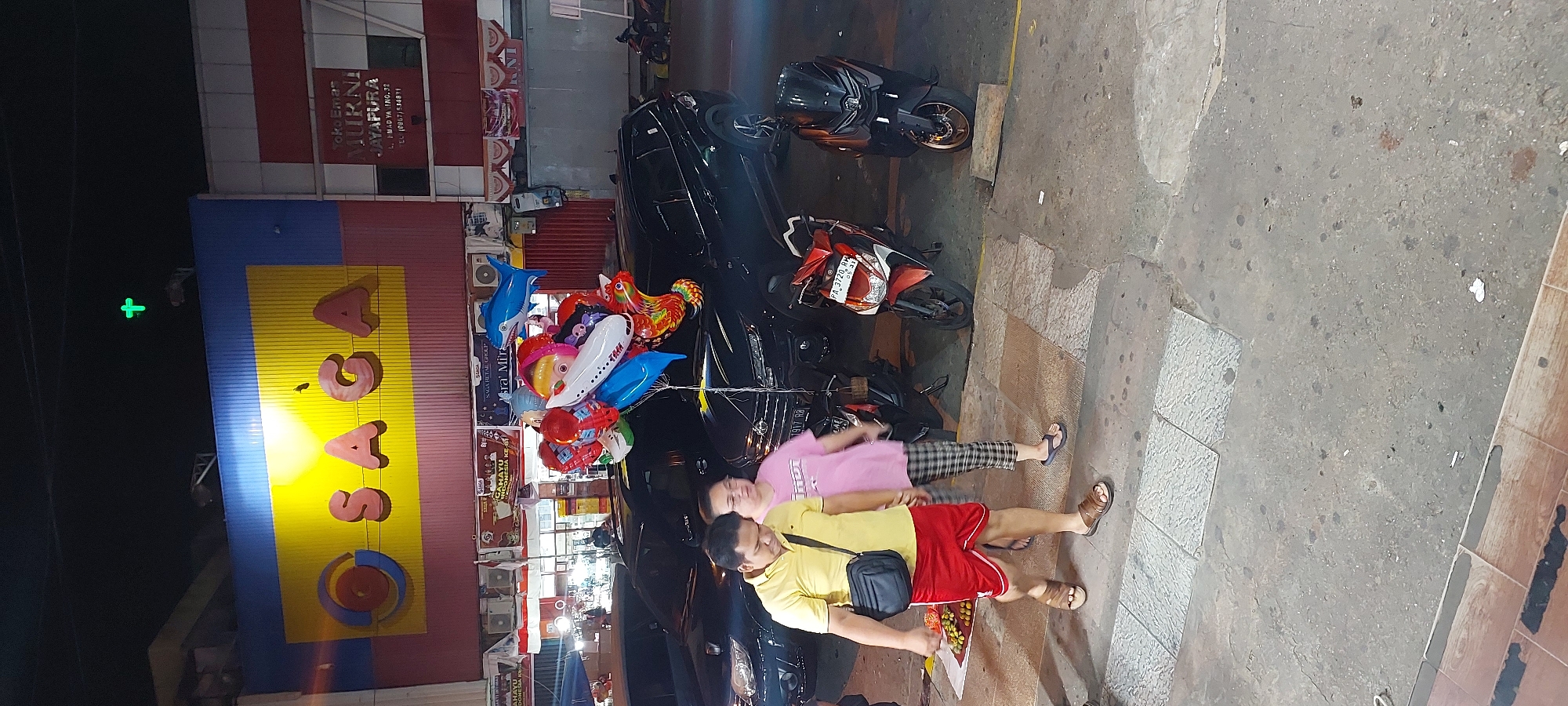 The image shows a street scene at night in Jayapura, Indonesia. A large building with a bright yellow sign that reads "SAGA" is visible in the background. The sign features a circular logo with red and blue rings. Above the "SAGA" sign, a green cross-shaped light is illuminated. To the right of the "SAGA" sign, there is another sign that reads "Toko Emas MURNI JAYAPURA" (Murni Gold Shop Jayapura).
In the foreground, two people are walking across a pedestrian crossing. The person on the left is a man wearing a yellow polo shirt, red shorts, and brown sandals. He has a black bag slung across his body. The person on the right is a woman wearing a pink t-shirt and checkered pants. She is wearing flip-flops.
Between the people and the building, several black SUVs are parked. In front of the SUVs, a cluster of colorful balloons are tied to a pole, including balloons shaped like a shark, an airplane, and a dragon.
Several motorcycles are parked on the right side of the image. The ground is paved with concrete and tiles. The overall scene appears to be a commercial area at night, possibly near a shopping center.