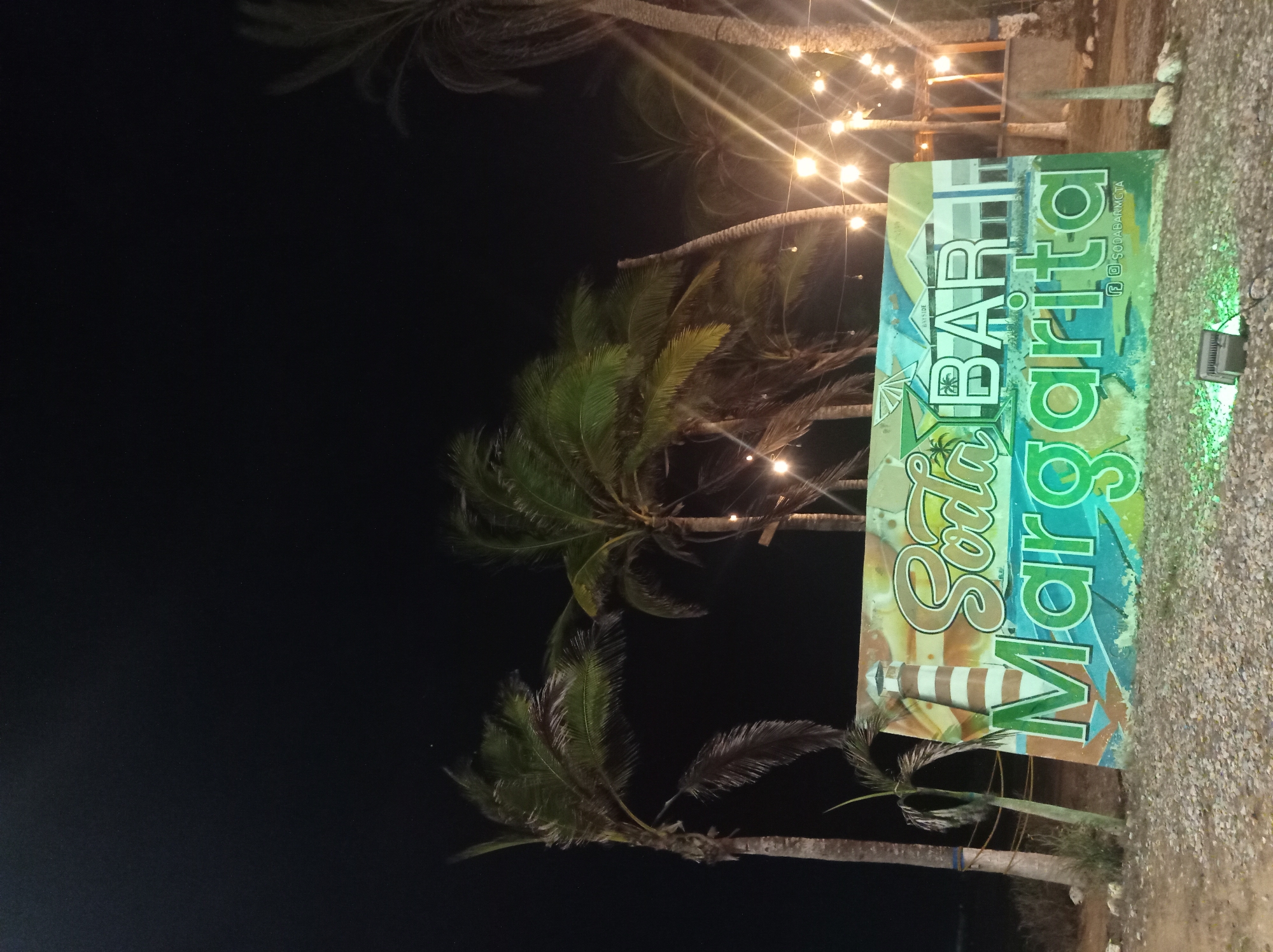 This image captures a vibrant outdoor bar scene at night, likely in Porlamar, Venezuela. The central subject is a large, colorful sign oriented vertically, featuring the name "Soda BAR Margarita" in stylized, flowing green and orange text. Below "BAR," a white and brown striped bottle graphic is depicted. Smaller text on the sign reads "BAYSIDE" and social media handles "f osodabarmgta," indicating Facebook and Instagram presence for "sodabarmgta."
The sign is positioned on what appears to be a gravel or crushed shell ground. To the right of the sign, a square light fixture emits a green glow onto the ground. The background is dominated by several tall palm trees with visible fronds, silhouetted against a dark, night sky. Strands of warm-toned string lights with bare bulbs are strung between the palm trees, casting a soft, inviting glow and creating light flares in the image. The overall setting suggests a relaxed, tropical, and festive atmosphere, typical of a beachfront or resort bar area designed for evening enjoyment. There are no people visible in the frame.