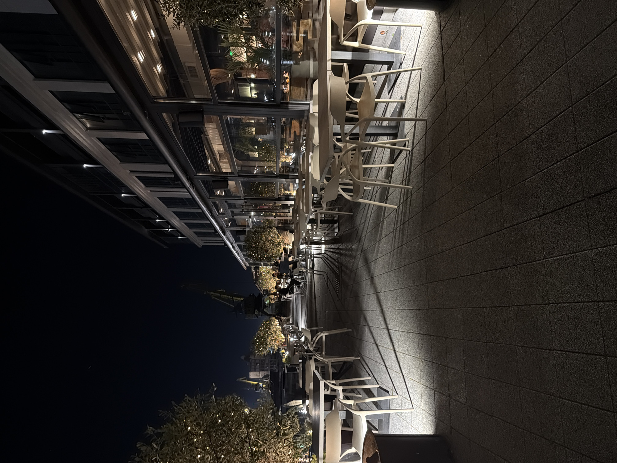 The image shows an outdoor restaurant patio in Burgas, Bulgaria at night. Rows of light-colored plastic chairs are stacked next to tables under a glass-enclosed canopy. The canopy reflects the lights and activity of the restaurant interior. Outside, there are streetlights illuminating trees. Cranes are visible in the distance, suggesting a port or industrial area. The patio surface appears to be tiled. The sky is dark, and the overall lighting is dim but artificial. No people are clearly visible in the foreground.