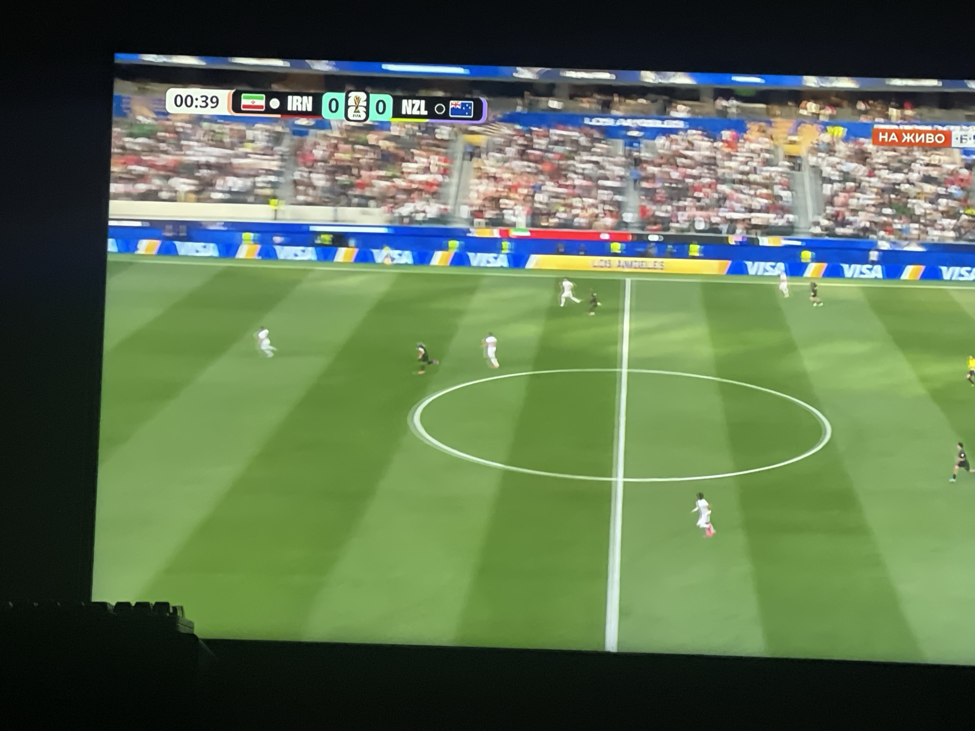 This is a photograph of a television screen displaying a soccer match. The screen shows a score graphic at the top indicating the time is 00:39 and the score is 0-0 between IRN and NZL. The field is green with white lines and a circle in the center. Several players in white and black uniforms are visible on the field. The background shows stadium seating filled with spectators, and banners advertising "VISA" and "LOS ANGELES." A red banner with Russian text "НА ЖИВО" (meaning "Live") is visible in the upper right corner of the stadium seating. The image is taken from an angle, with a portion of a black keyboard visible in the lower left corner. There is no indication that this image is located in the city of Ruse, Bulgaria.