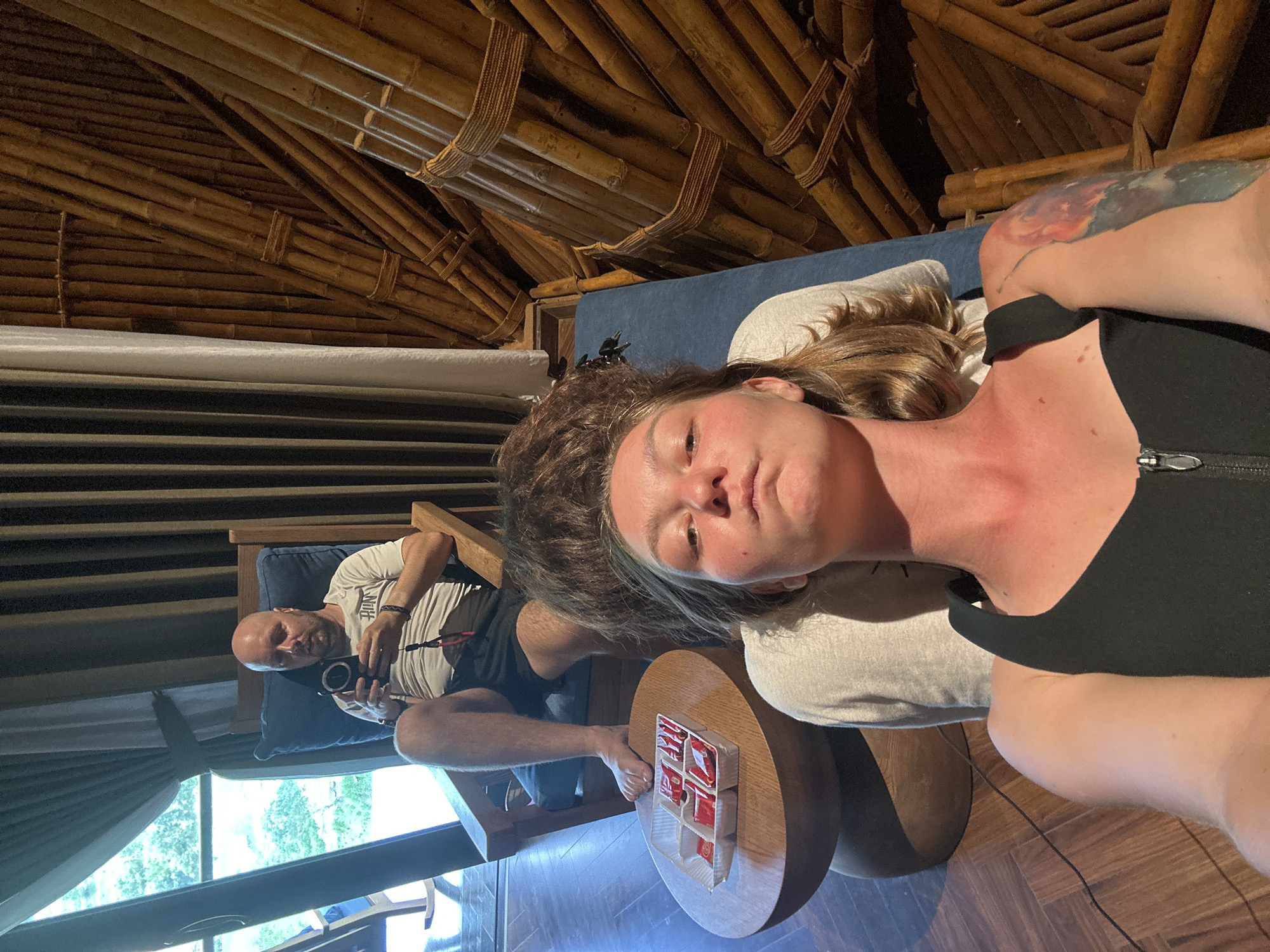 This indoor scene, located in Mù Cang Chải, Vietnam, depicts two individuals within a well-lit room featuring both modern and rustic elements.
In the foreground, a woman lies on her back on a blue cushioned surface with a white pillow, engaged in taking a selfie. She has brown hair and wears a black, zippered tank top. A colorful tattoo is visible on her left upper arm. Her face, neck, and chest exhibit areas of redness, consistent with sun exposure. To her left, a round wooden table holds a clear plastic container segmented into six compartments, each containing a red-wrapped item. A set of keys is placed on the blue cushion near her head.
In the mid-ground, a bald man with a beard sits in a wooden rocking chair equipped with blue cushions. He wears a light-colored t-shirt with "RUN" inscribed across the chest and dark shorts, his feet bare. His gaze is directed downwards at a black handheld electronic device. Behind him, a large window or glass door on the left side of the room offers a bright outdoor view of green foliage. White curtains partially frame this opening.
The right wall of the room is distinctively constructed from numerous bamboo poles, arranged vertically and secured with rope bindings, providing a textured, natural aesthetic. The floor is composed of light-colored wooden planks. A black electrical cable is visible on the floor, extending from the area of the round table. No direct interaction between the two individuals is observed.