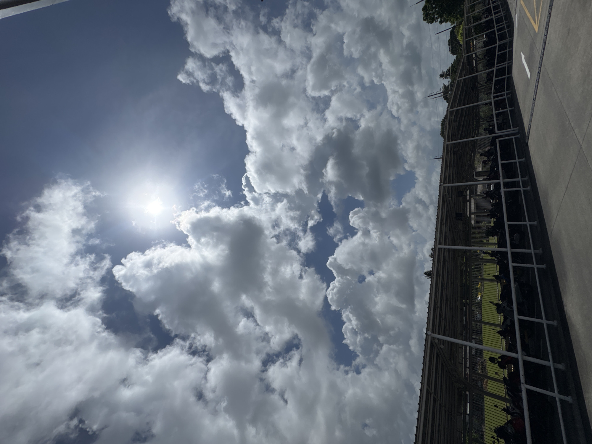 A wide-angle, full shot captures a covered parking area in Chalong, Thailand, under a partly cloudy sky with the sun prominently visible. The parking area is elongated, featuring a white metal roof supported by numerous white poles, and a white metal railing runs along the front of the covered space. Parked beneath the shelter are rows of motorcycles, many of them dark-colored. A concrete pathway is in the foreground, marked with a white arrow pointing forward and yellow lines to its right. Beyond the parking area, a green grassy field is visible, with trees and utility poles in the background. The sky is a mixture of deep blue and bright white clouds, with the sun creating a bright glare.