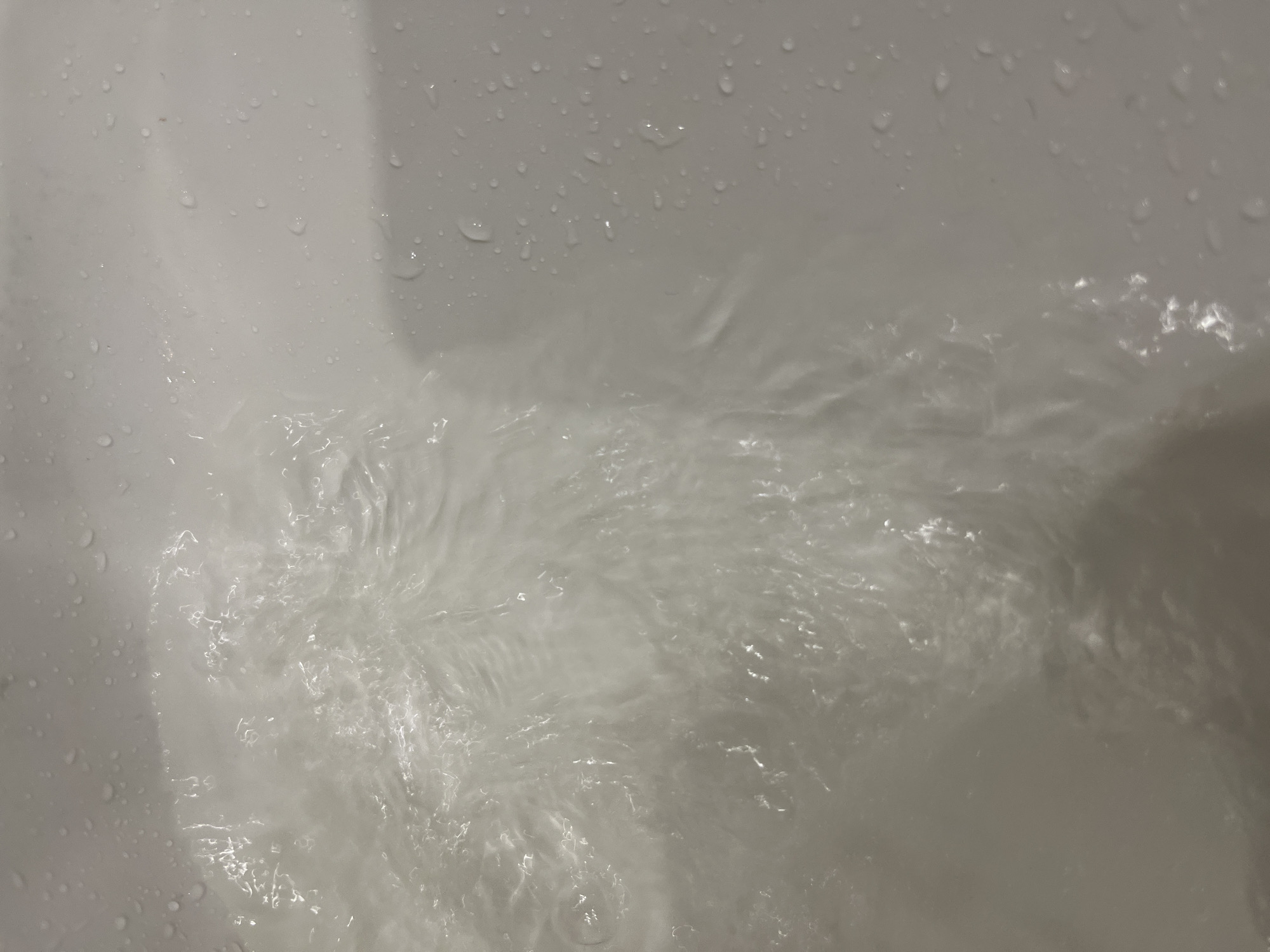 A close-up, overhead shot captures the swirling surface of water in a white sink or bathtub. The water appears clear and is in motion, with ripples and reflections creating a dynamic, textured pattern. Small droplets of water are visible on the surrounding white surface, suggesting a damp environment. The lighting is somewhat dim, creating soft shadows and highlights on the water and the surface. The scene is devoid of people or any identifiable objects other than the water and the basin itself. The location is unknown.