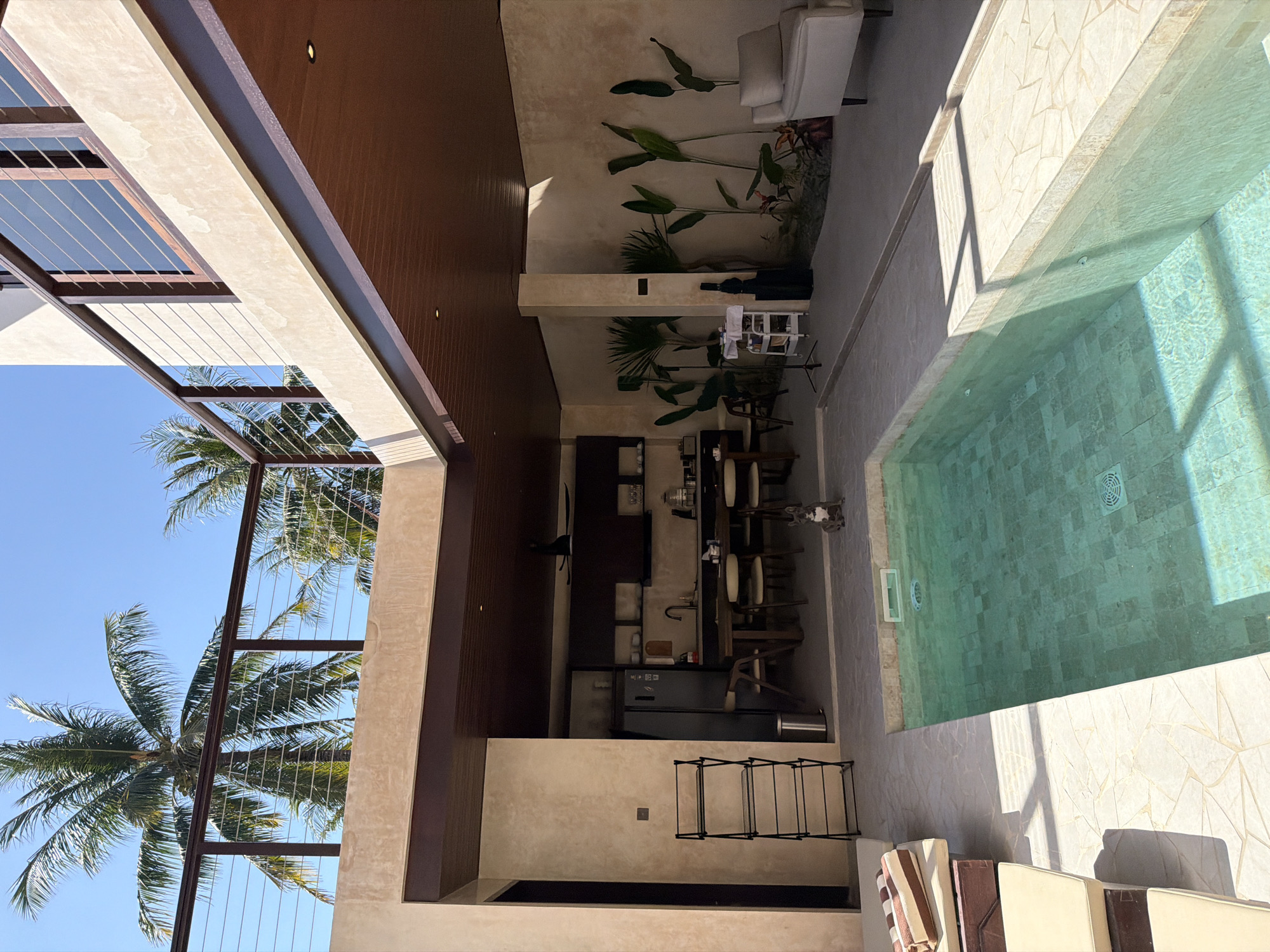 In Kuta, Indonesia, this image captures a private outdoor living space adjacent to a swimming pool. The scene features a small, rectangular pool with turquoise water, tiled in a mosaic pattern. Surrounding the pool is a light-colored tiled patio area. To the left, a section of a wooden lounge chair with striped cushions is visible.
Behind the patio, an open-air kitchen and dining area are set up. This area has a dark wood ceiling and textured beige walls. A dark refrigerator, wooden dining table with chairs, and kitchen counter with shelves are present. To the right of the kitchen area, an outdoor space is furnished with potted tropical plants, a white trolley cart, and a dark case leaning against a pillar. Further to the right, a light-colored armchair sits on a tiled floor.
Overhead, a balcony with a dark railing and cable wires is visible, supported by wooden beams. Palm trees with lush green fronds and a clear blue sky form the background. A small grey and white French bulldog is sitting on the patio near the pool's edge. The overall impression is of a modern, open-plan dwelling in a tropical setting.