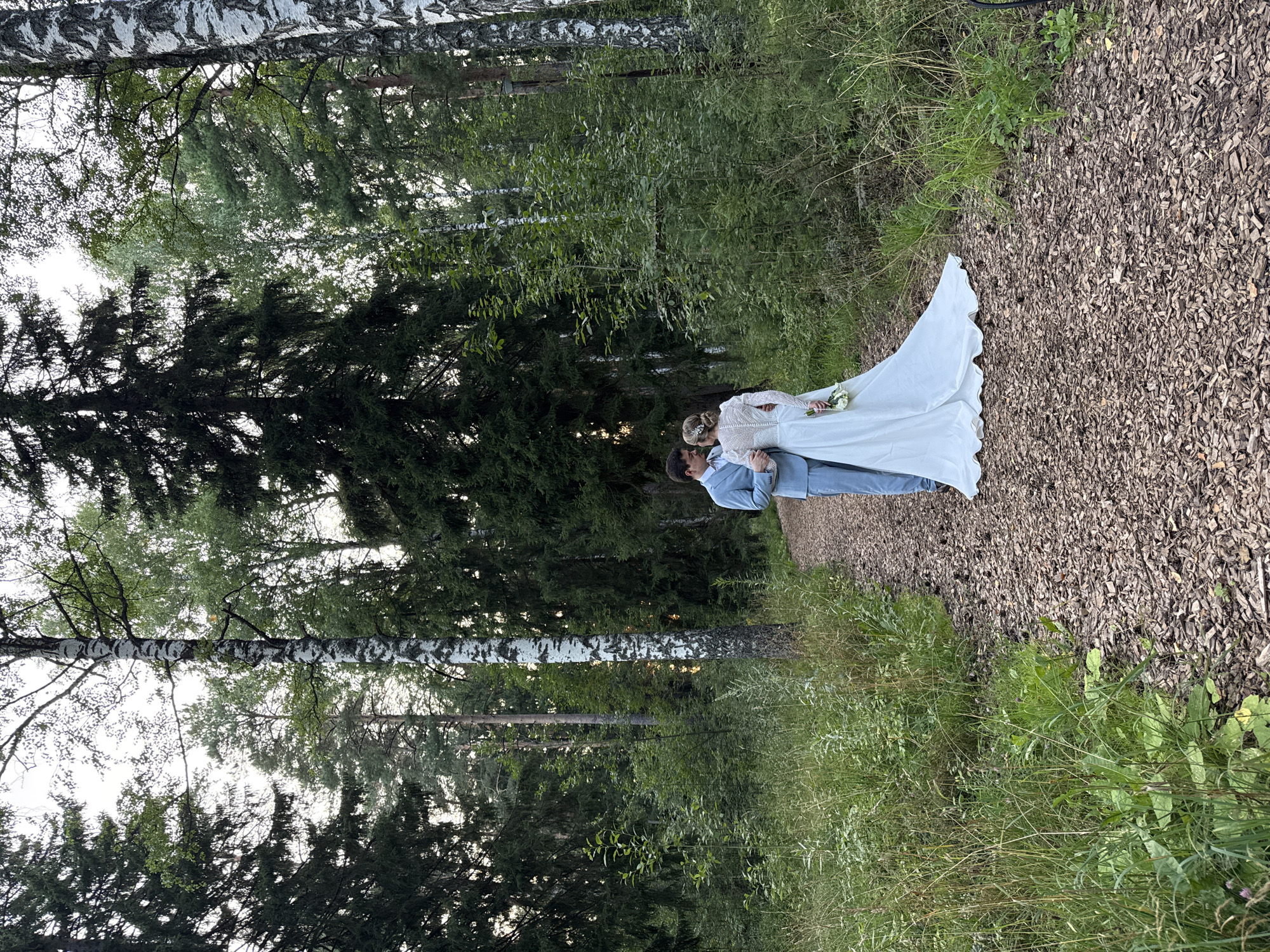 A newly married couple stands on a wood chip path in a dense forest in Bakhtiyary, Russia. The bride, wearing a long-sleeved white wedding dress and holding a bouquet, faces her groom. He is dressed in a light blue suit and has his arm around her. The path is surrounded by lush green trees and foliage, with tall birch and pine trees dominating the landscape. The lighting suggests it is late afternoon or early evening, with soft natural light filtering through the trees.