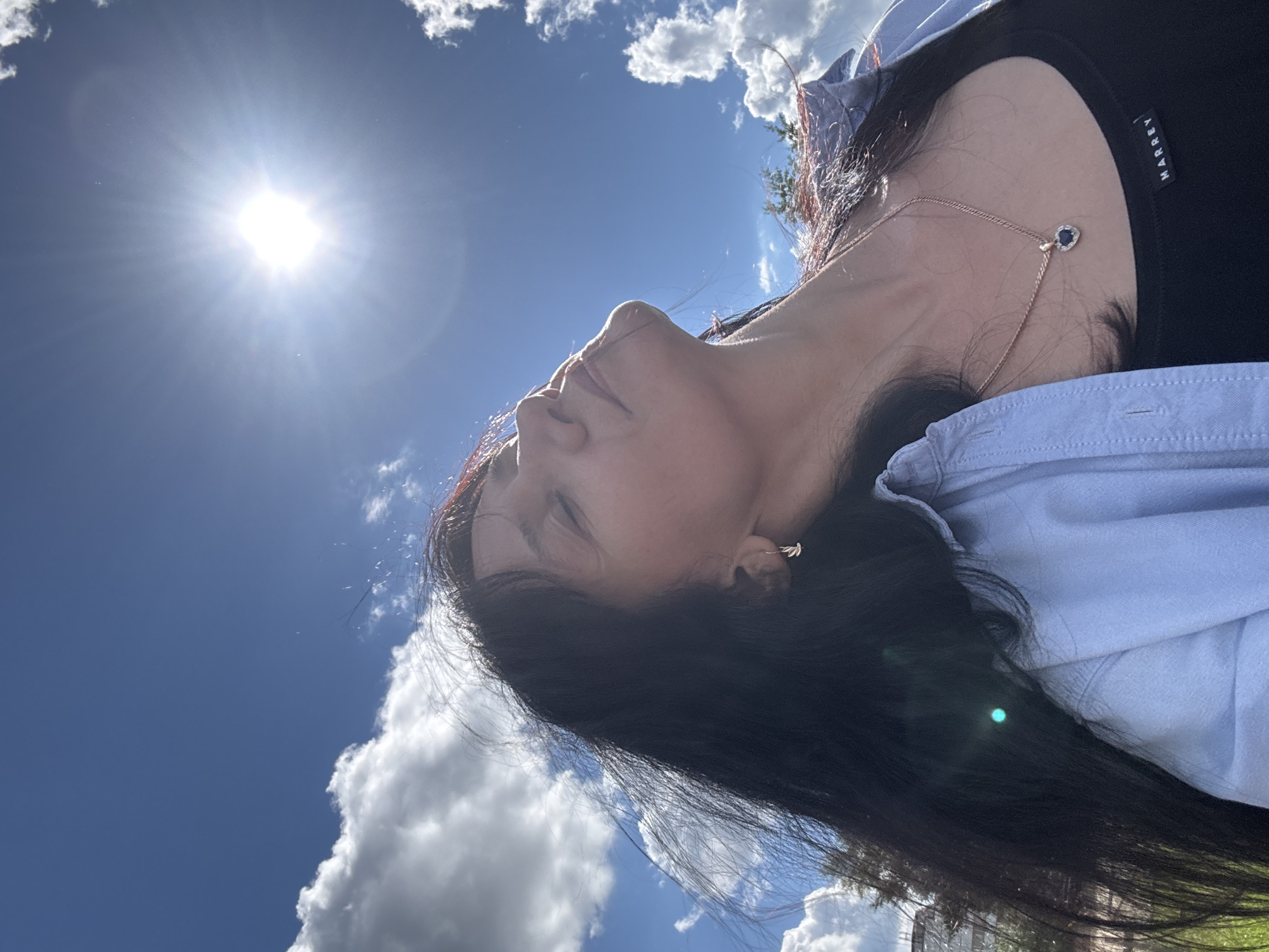 An outdoor, low-angle, medium shot shows a woman with long, dark hair tilting her head back towards a bright sun against a blue sky with scattered white clouds. She is wearing a light blue collared shirt over a black tank top and a delicate rose gold chain with a heart-shaped blue pendant. Her eyes are closed and her face is tilted upwards, suggesting she is enjoying the sun. The sun is high in the sky, creating lens flare and beaming rays. The background shows a slightly out-of-focus green grassy area with some trees and buildings visible in the distance, hinting at a park or open space within the city of Chaikovsky, Russia.