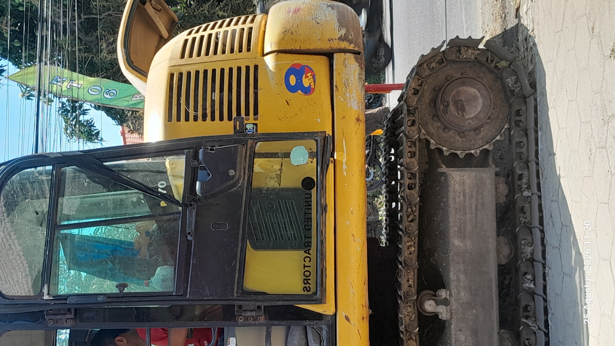 This is a close-up, medium-angle, eye-level photograph of a yellow excavator in Jomblang, Indonesia. The excavator is positioned on a light-colored paved surface, likely a sidewalk, with a road visible in the background. A portion of the excavator's track is prominent in the foreground. The excavator's cabin, with tinted windows, occupies the left side of the frame. A person wearing a red shirt is visible inside the cabin, appearing to operate the machinery. A reflective sticker with a blue "8" and the word "DASH" is affixed to the yellow body of the excavator on the right side. A green flag with some writing on it is partially visible in the upper right quadrant of the image, suspended above the excavator. The environment suggests an outdoor setting with daylight illumination. The bottom left corner of the image displays a timestamp: "25 Agustus 2026 11:20."