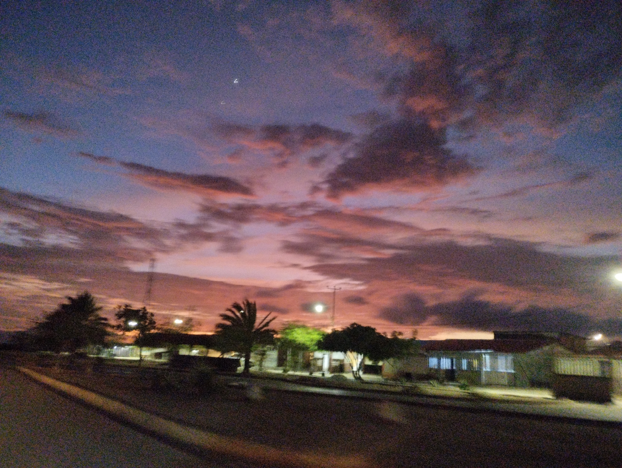 The image captures a dusky sky over a residential area in El Espinal, Venezuela. The sky is a gradient of deep blue and purple, with streaks of pink and orange clouds reflecting the setting sun. A few bright points, possibly stars or distant lights, are visible in the upper part of the sky. In the foreground, a dark, paved road or pathway stretches into the distance. To the left and right of the road are low-lying buildings, appearing to be houses, with lights on inside some of them, suggesting it is dusk or early evening. There are scattered trees and vegetation around the buildings. A utility pole with some wires is visible behind the buildings. The overall scene depicts a quiet, rural or suburban setting at twilight.