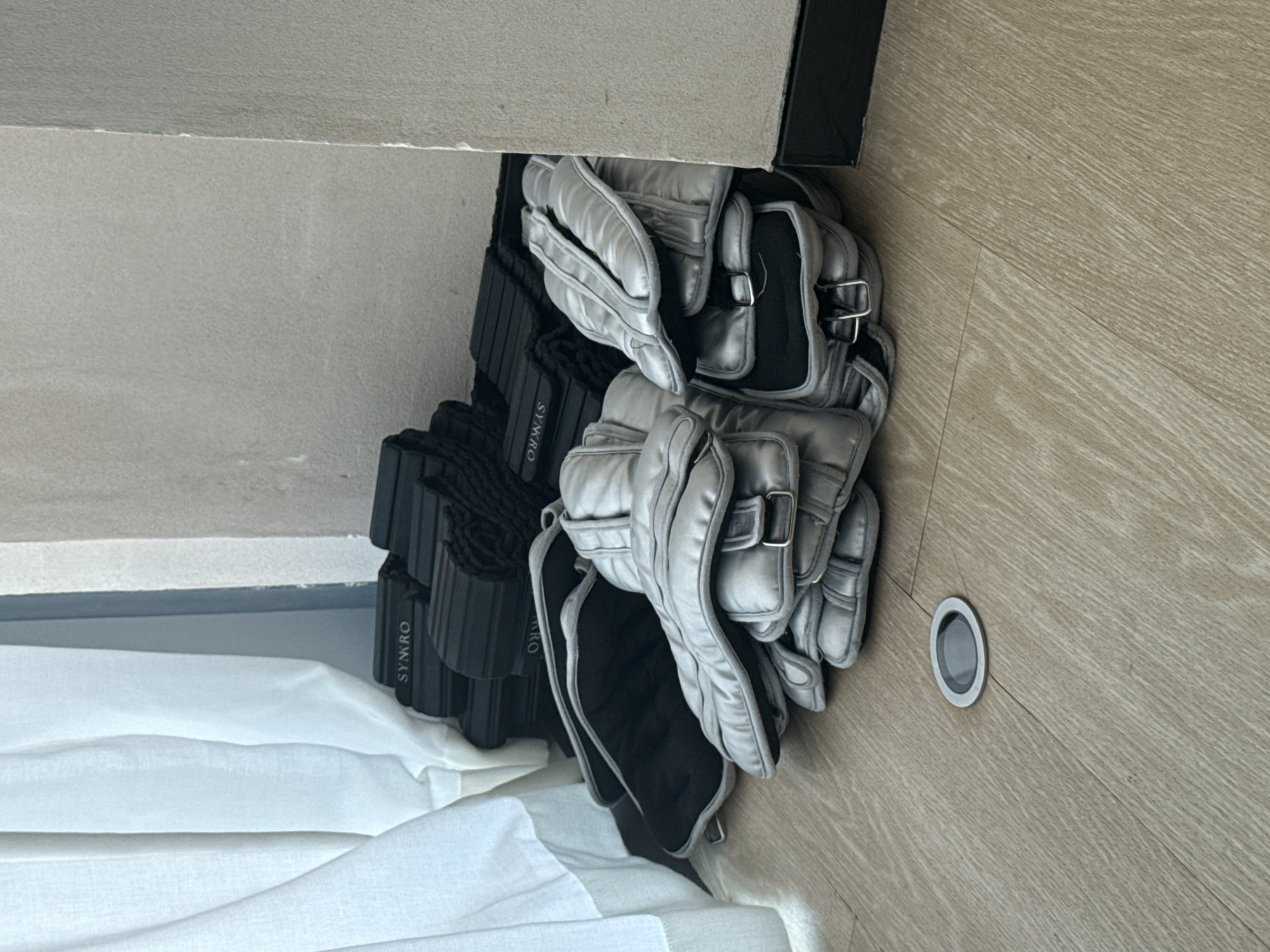 A pile of black yoga blocks with the logo "SYNRO" is stacked against a light gray wall. In front of the yoga blocks, several pairs of silver weight-lifting gloves with black straps and buckles are arranged in a heap on a light-colored wooden floor. A white curtain is visible on the left side of the image, partially obscuring the wall. A small, circular, metallic object is embedded in the floor in the lower left quadrant. The scene appears to be indoors, likely in a gym or fitness studio, possibly located in Dubai, United Arab Emirates. There are no people or animals present in the image, and no action is depicted.