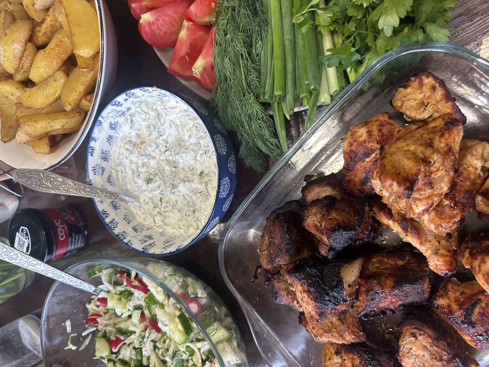 A high-angle shot captures a spread of food arranged on a dark surface, likely a table, in Chaikovsky, Russia.
At the top left, a white bowl overflows with golden-brown potato wedges. Beside it, a plate showcases sliced tomatoes alongside fresh dill and green onions. A blue-and-white patterned bowl contains a creamy white sauce with green flecks, accompanied by a silver serving spoon.
A clear glass bowl in the lower left holds a salad composed of shredded cabbage, cucumber, radish, and dill, also with a silver serving spoon. To the right, a large clear glass baking dish is filled with pieces of grilled meat, some with darker, charred edges.
Between the salad and the patterned bowl, a small black jar with a visible label is present. In the background, fresh parsley sits beside the green onions.
The overall arrangement suggests a meal setting, possibly a gathering or a family dinner, showcasing a variety of dishes.