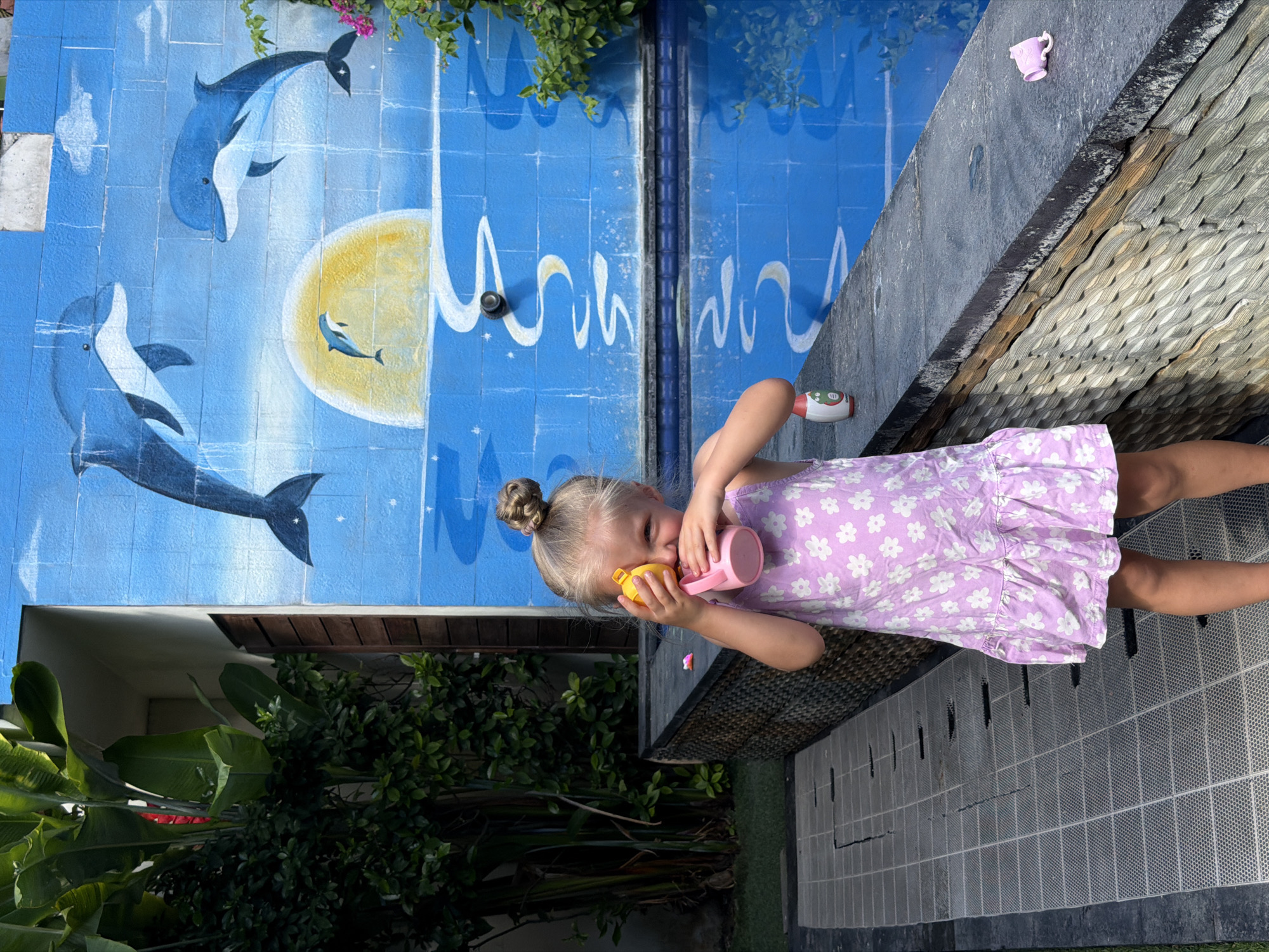 A young Caucasian girl, with blonde hair in a bun, wearing a purple floral dress, stands by a swimming pool in Kuta, Indonesia. She is holding a pink cup to her face, appearing to drink from it. The background features a mural of dolphins and a sun on a blue brick wall. Lush green foliage is visible to the left of the girl. The swimming pool is blue and reflects the mural. The ground in front of the girl is a grey tiled area. A small pink toy cup is visible on the edge of the pool to the right of the girl.
