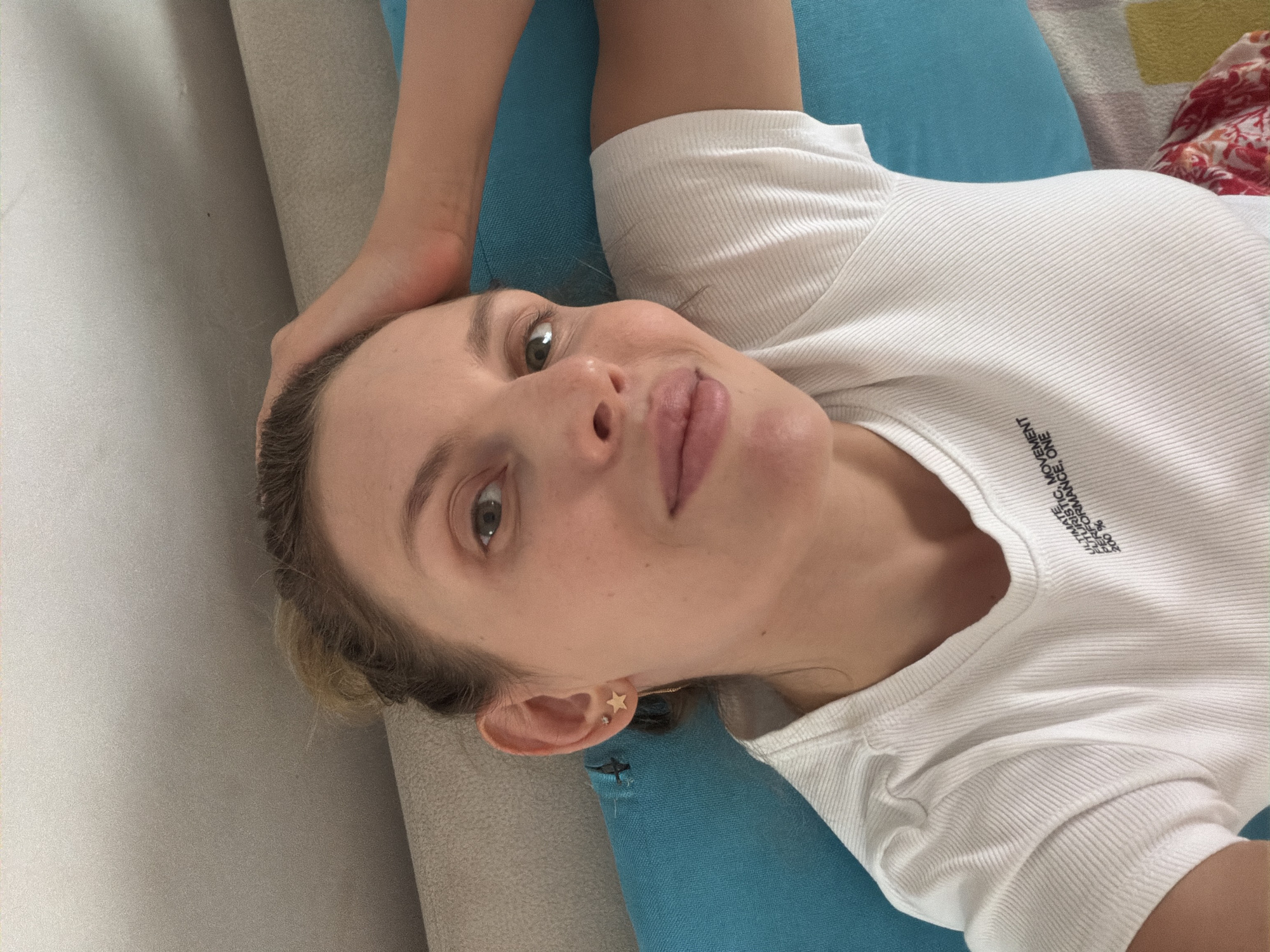 The image shows a young woman lying on her side on a blue cushion, with her head resting on a grey couch. She is wearing a white t-shirt with black text on the front. Her hair is pulled back in a bun. Her right hand is raised and resting on her head. She is looking towards the camera with a slight smile. The background appears to be an indoor setting, possibly a living room or bedroom, with a patterned blanket visible at the bottom right of the frame. There are no specific landmarks or details in the image that would allow for the precise identification of the location as Budva, Montenegro.