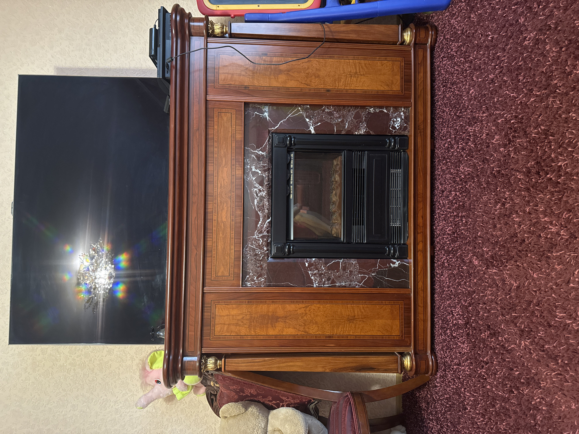 The image shows a large, dark brown wooden fireplace with a black insert. Above the fireplace is a large, black flat-screen television. The television screen displays a reflection of a chandelier, with rainbow-colored light refracting from it. To the left of the fireplace is a portion of a plush, burgundy armchair with a light brown teddy bear sitting on it. To the right of the fireplace, a blue plastic toy structure is visible. The fireplace is situated on a shaggy, deep red carpet. The background consists of a textured, light-colored wallpaper. The scene appears to be an indoor living space.