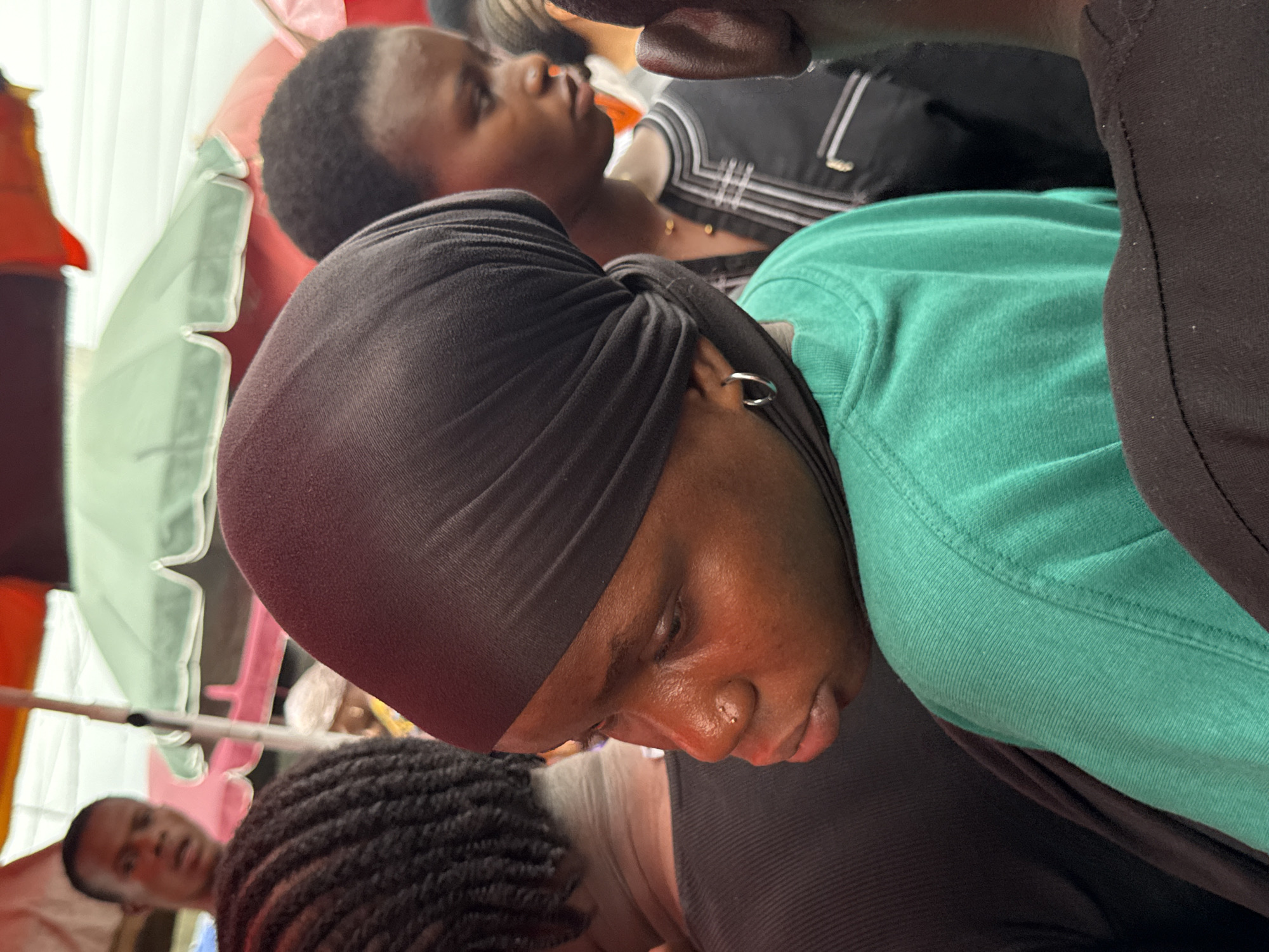 The image shows a close-up, angled view of several individuals in what appears to be a crowded outdoor setting, possibly a market or public gathering. The main focus is on a person wearing a dark head covering, a green shirt, and a dark outer garment. They have a piercing in their ear and what seems to be a piercing on their nose. The person has their head down, and they are looking downward.
Behind them is another person facing away and wearing a dark top with white stripes and gold-colored details, while another person is looking up in the background. Other individuals and objects are partially visible, including a red-and-white striped awning or umbrella, and another individual in the bottom left of the frame. Due to the angle and limited field of view, details about the wider environment are not clear, making it difficult to confirm the exact location in Lekki, Nigeria or the nature of the event.