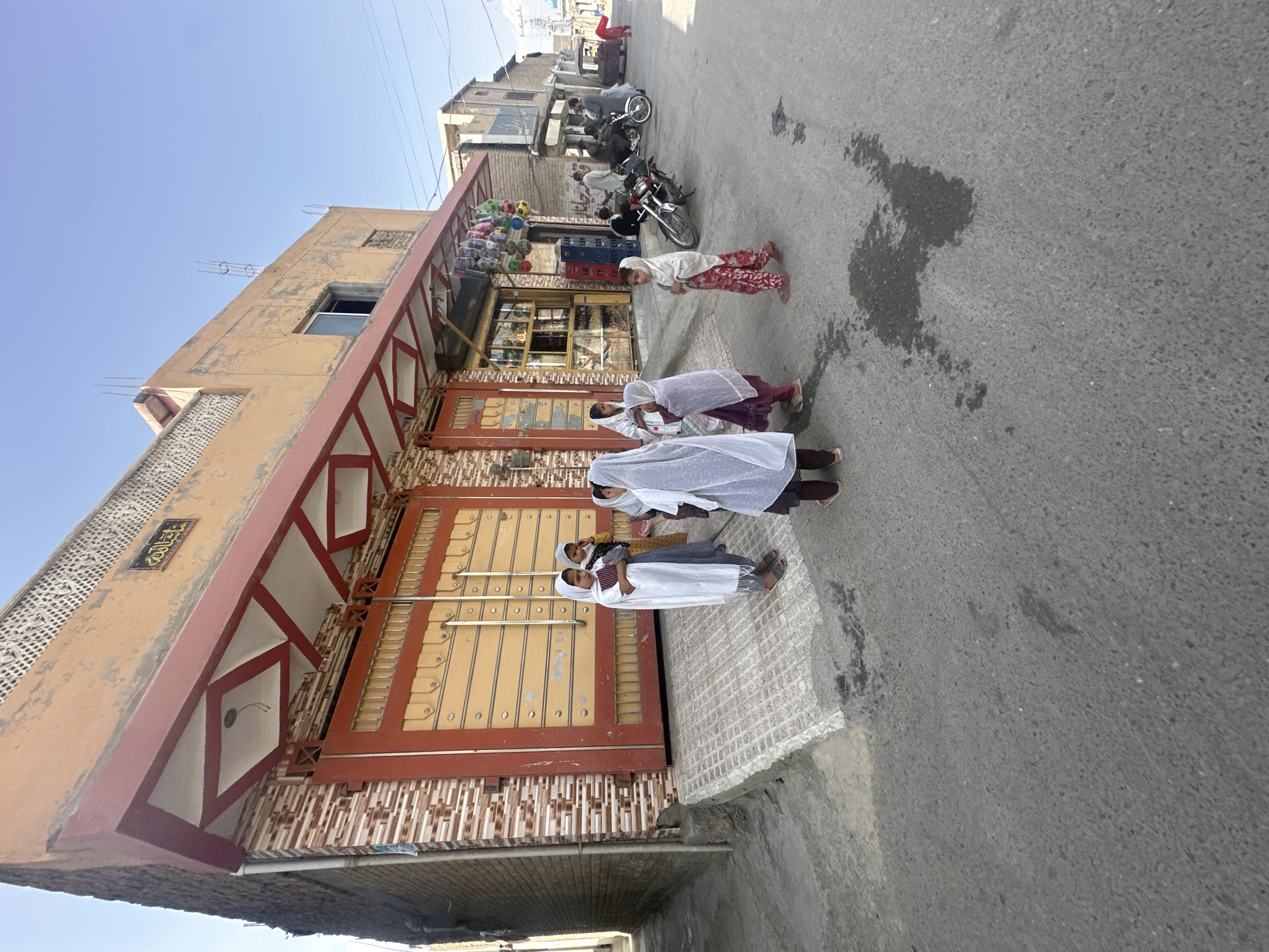 The image depicts a street scene in Quetta City Tehsil, Pakistan. A group of four girls stands in front of a building with a large, gated entrance. The girls appear to be wearing traditional clothing, with white headscarves. The building is two stories tall and made of light-colored brick. It has a brown-and-red gate. A small shop is visible nearby, selling various items. There are a few motorbikes are parked along the street. The street itself appears to be paved and slightly wet. The sky is clear and blue.