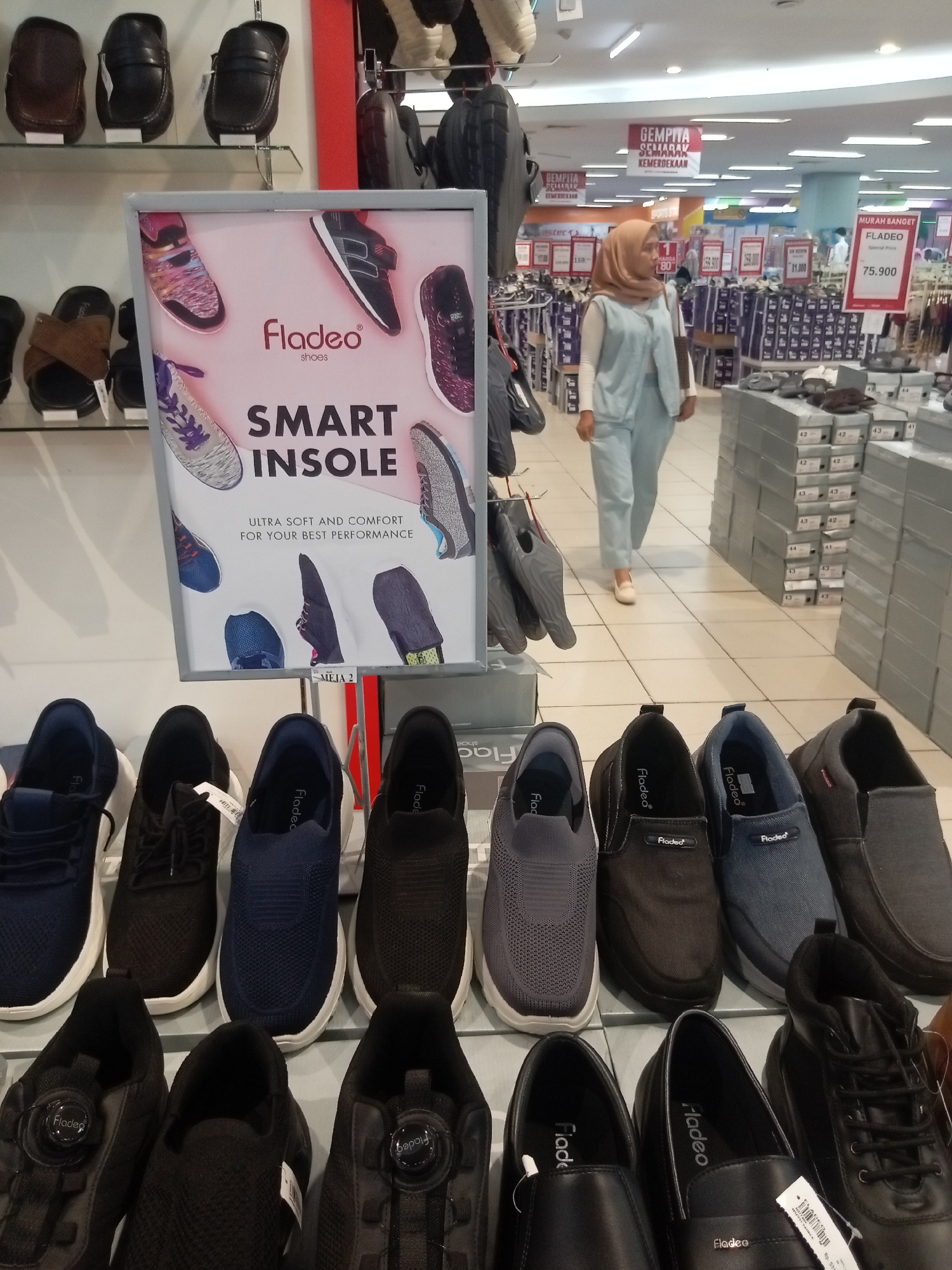 This image depicts the interior of a shoe store in Batam, Indonesia, likely a department store or a standalone shoe retailer. The display features various types of footwear, including loafers, sneakers, and casual shoes, arranged on shelves and in cardboard boxes. A prominent sign advertises "Fladeo Shoes" with the slogan "SMART INSOLE" and describes the product as "ULTRA SOFT AND COMFORT FOR YOUR BEST PERFORMANCE." In the background, a woman wearing a light blue hijab and matching pantsuit walks past shoe displays. Other shoppers and store employees are visible in the blurred background, and promotional signage for "GEMPITA SEMARAK KEMERDEKAAN" (Independence Day Celebration) suggests the photo was taken around the Indonesian Independence Day. The overall environment is a well-lit retail space with multiple rows of shoe displays and stacked boxes of merchandise.