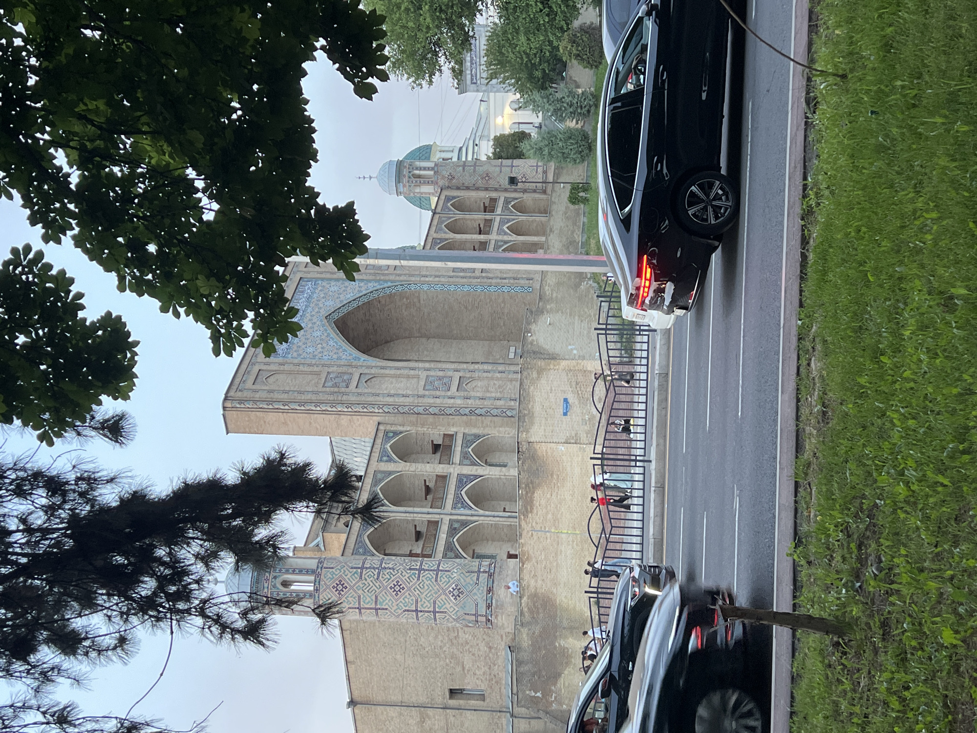 This image shows a street scene in Tashkent, Uzbekistan, featuring the Khast Imam complex. The architectural structures, characterized by their intricate tilework and domes, are partially obscured by trees and vehicles. The street, divided by lane markers, has pedestrian traffic along the metal railing. There are at least two black vehicles on the right side of the frame, one is stopped with its brake lights illuminated. The sky is overcast, suggesting a dull or cloudy day.