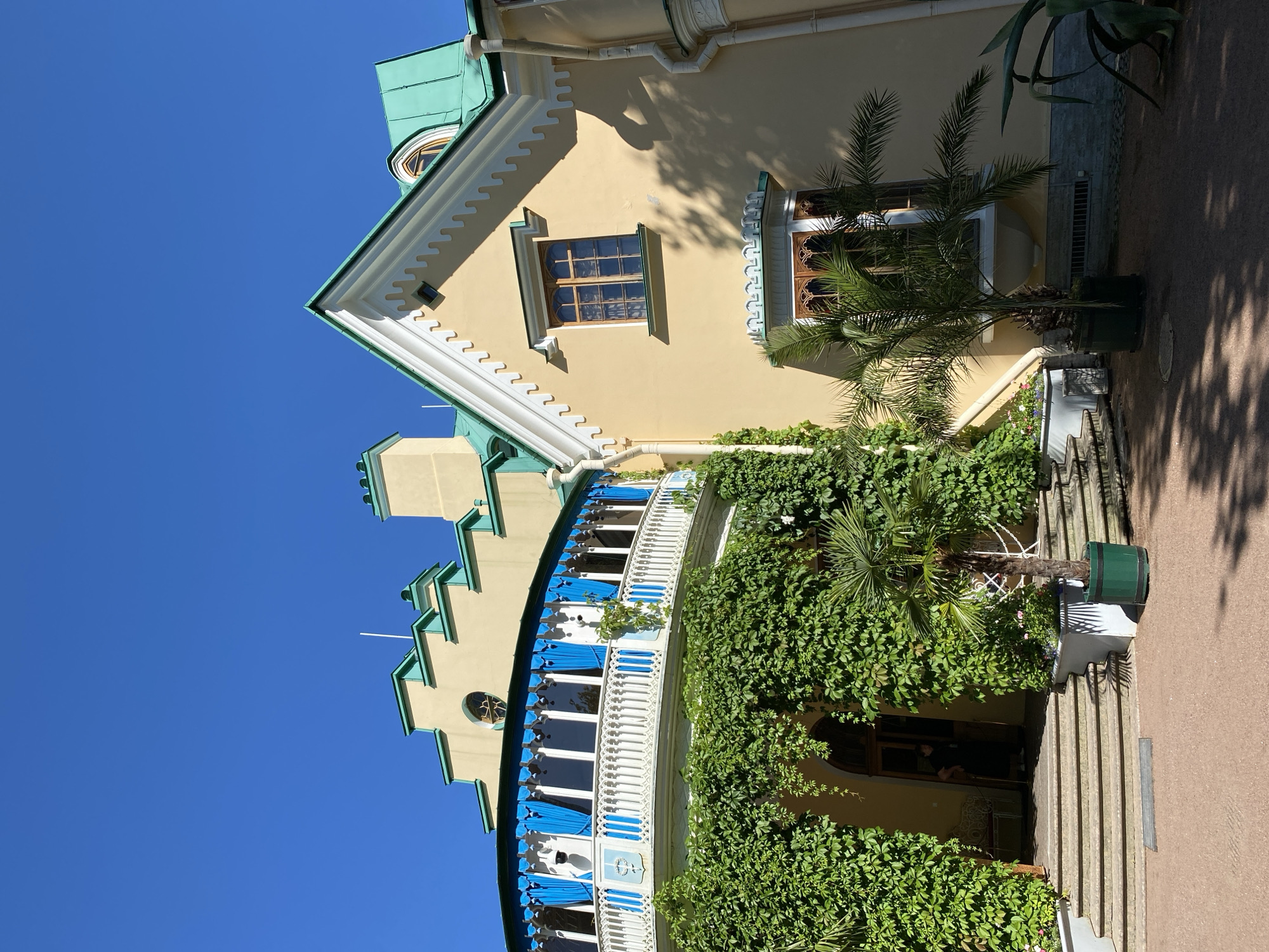 The image captures a section of a yellow building with a green roof, set against a clear blue sky. The building features a curved balcony with white railings and blue curtains, partially covered by lush green ivy. A staircase leads up to an arched entranceway. Palm trees and other greenery are visible in the foreground. The architectural style appears to be a blend of various influences, possibly Gothic Revival or Moorish. This building is located within the grounds of the city of Peterhof, Russia, a suburban town in the federal city of Saint Petersburg, Russia.