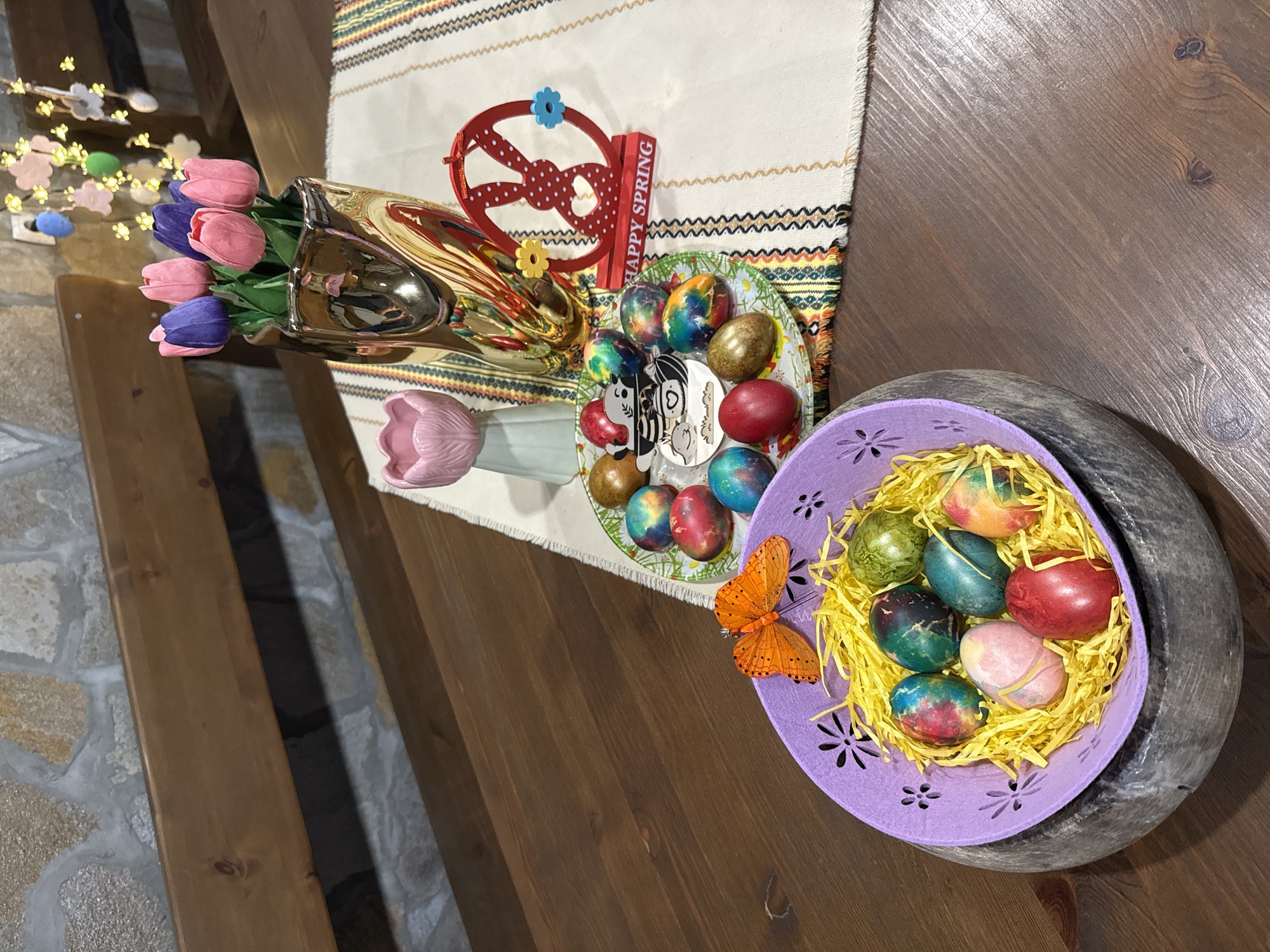 The image shows a table decorated for Easter, possibly in Lukovit, Bulgaria. A bouquet of pink and purple tulips sits in a gold vase, next to a decorative "Happy Spring" sign and a red bunny ornament on a table runner. A plate of colorful Easter eggs is nearby, accompanied by a purple basket filled with more decorated eggs and artificial yellow straw. An orange butterfly decoration adorns the basket. The table appears to be made of dark wood and is placed against a stone wall. A string of lights with decorative eggs is visible in the background.