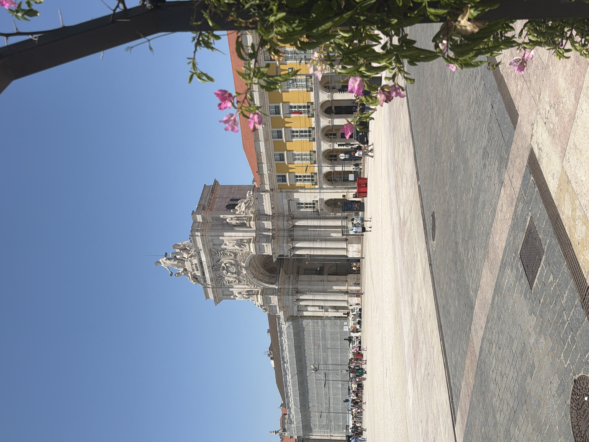 The image depicts a sunny day in Lisbon, Portugal, specifically at the Praça do Comércio. A grand triumphal arch, the Arco da Rua Augusta, dominates the background, adorned with intricate sculptures and topped with a statue. To the right of the arch, a yellow building with red-tiled roofs and arched balconies stretches across the square. The foreground features a vast, open cobblestone plaza, populated by numerous tourists and locals engaged in various activities such as walking and posing for pictures. Some are gathered near food stalls and shops. To the right, a trellised structure with pink flowers and green foliage partially obscures the view, adding a touch of natural beauty to the urban landscape. The sky is a clear, vibrant blue.