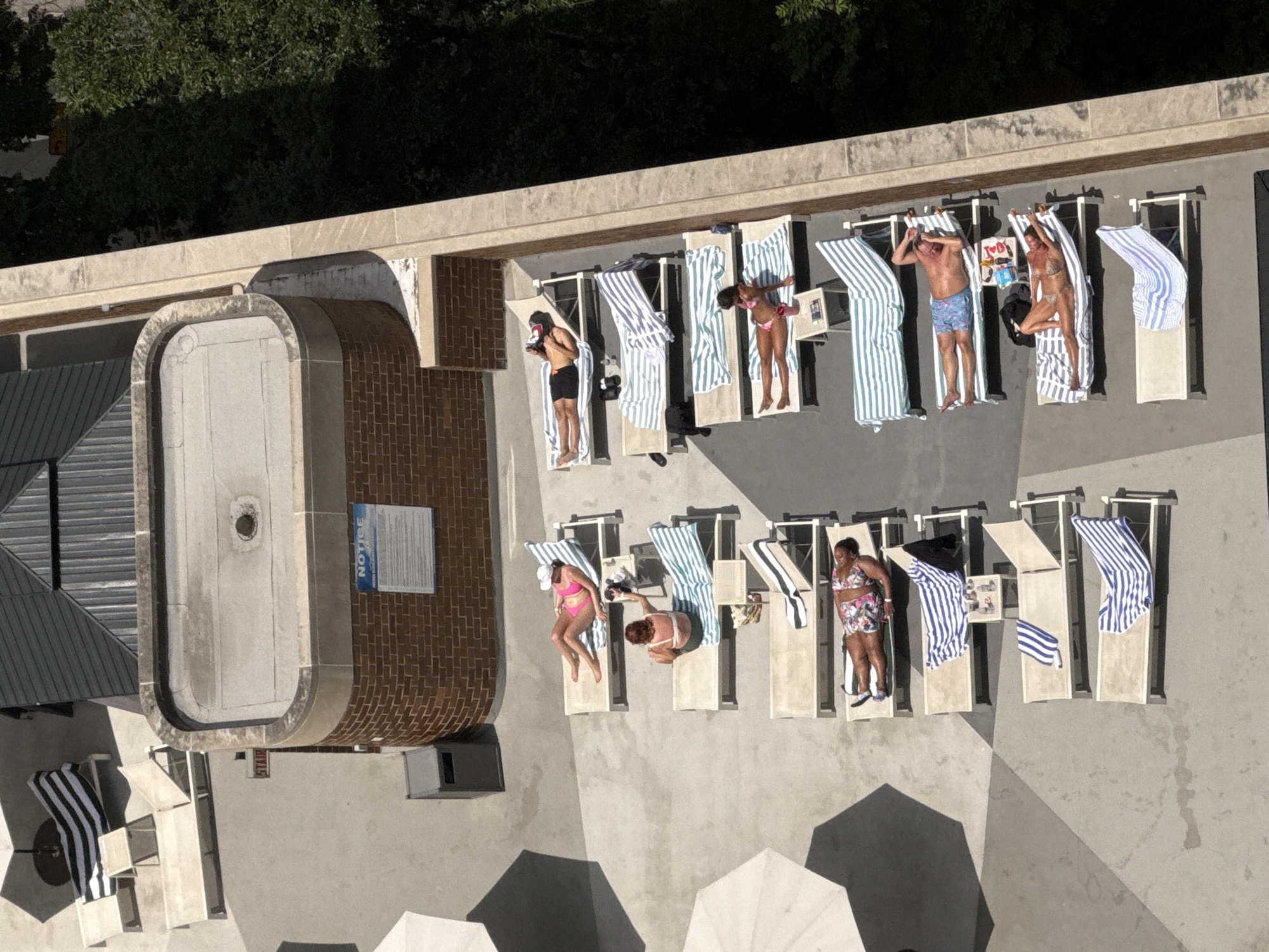 This is an aerial, high-angle, full shot of a rooftop deck in Chicago, Illinois, USA, on a sunny day. The deck is lined with lounge chairs, many of which are occupied by people sunbathing. Striped towels are draped over several chairs. In the foreground, a few people are visible, and further back, more lounge chairs are set up. Some of the chairs are empty. The background features trees and a brick building with a flat, grey roof. There is also a sign that says "NOTICE" attached to the brick building.
