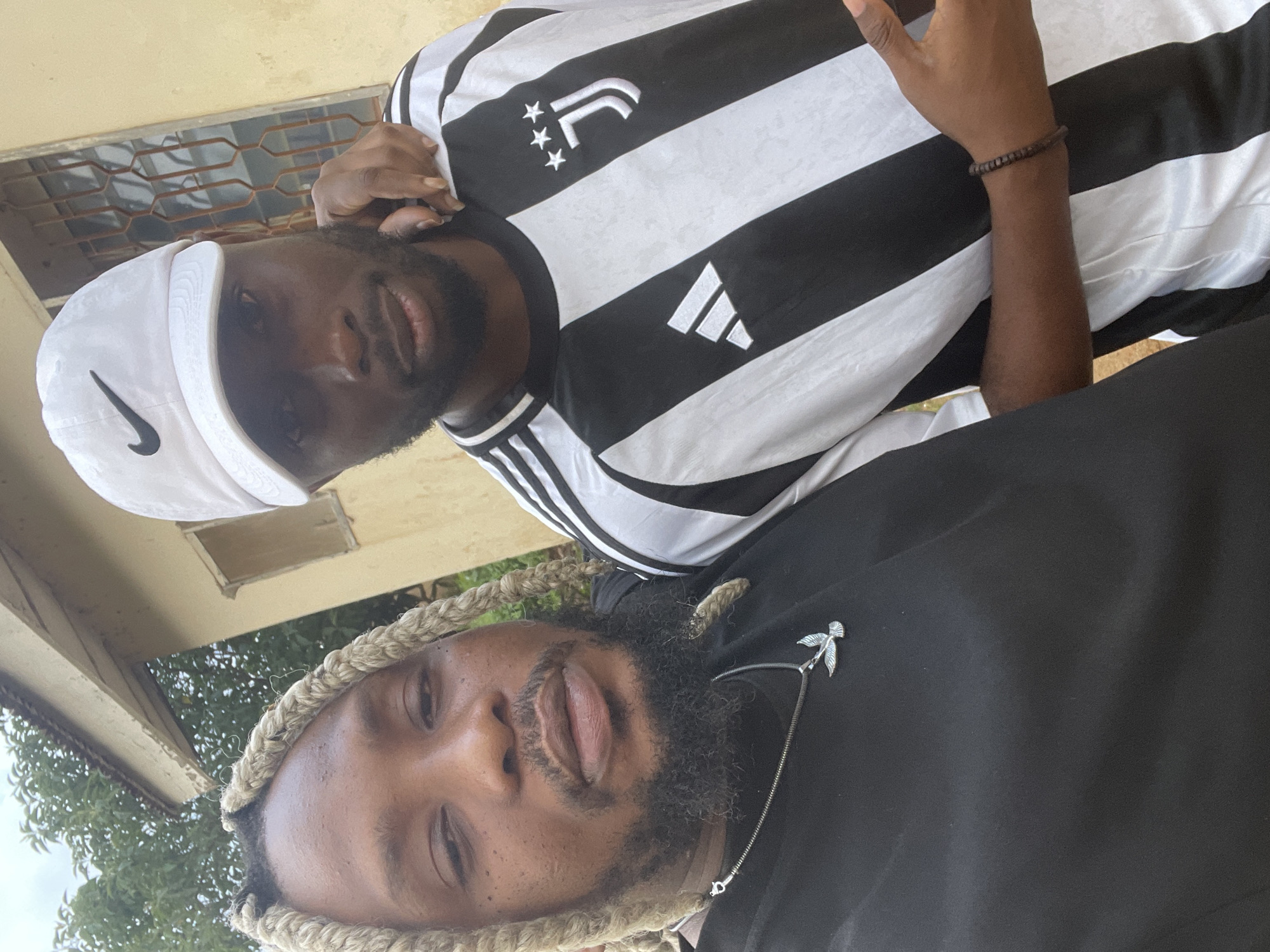 Two men are posing for a selfie outdoors. The man on the left has blonde braided hair and a beard, wearing a black t-shirt and a necklace with a bird pendant. The man on the right wears a white baseball cap and a black and white striped Juventus jersey, with a beaded bracelet on his wrist. His left arm is around the shoulder of the man on the left. They are standing in front of a building with a yellow facade and a window with metal bars. Trees are visible in the background. The overall impression is that of a casual, possibly urban outdoor setting. Based on the presence of certain architectural and environmental cues that are common in Jos, Nigeria, it is likely this scene is located within the city of Jos, Nigeria.