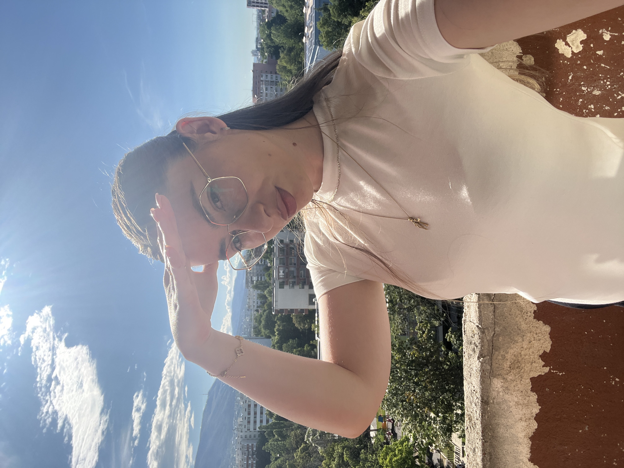 A medium shot captures a woman with long dark hair, wearing glasses and a white t-shirt, standing on a balcony with a city view under a bright sky. She is looking towards the right, her right hand shielding her eyes from the sun. The background features buildings, trees, and mountains in the distance. The balcony railing in the foreground is weathered and shows signs of age.