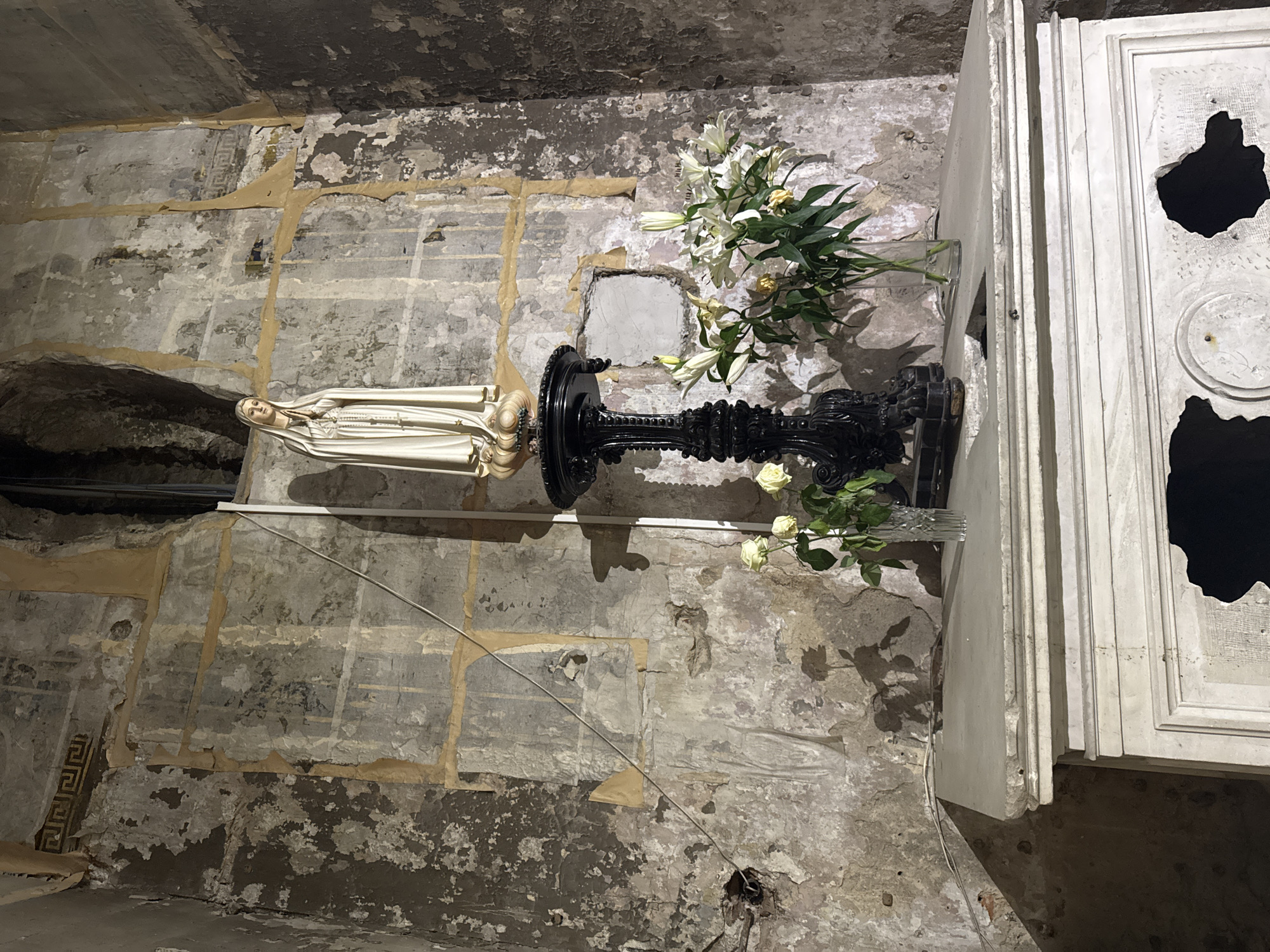 The image shows an indoor space, possibly a crypt or a damaged religious space, in Saint Petersburg, Russia. A white statue of the Virgin Mary is positioned on a black, ornate pedestal. A vase of lilies and white roses sits near the statue. The surrounding walls are severely damaged with peeling paint and exposed layers, hinting at age and neglect. A white, heavily damaged cabinet or architectural element with large holes is also present. Electrical wiring is visible against the wall. The scene is dimly lit, contributing to a somber and neglected atmosphere.