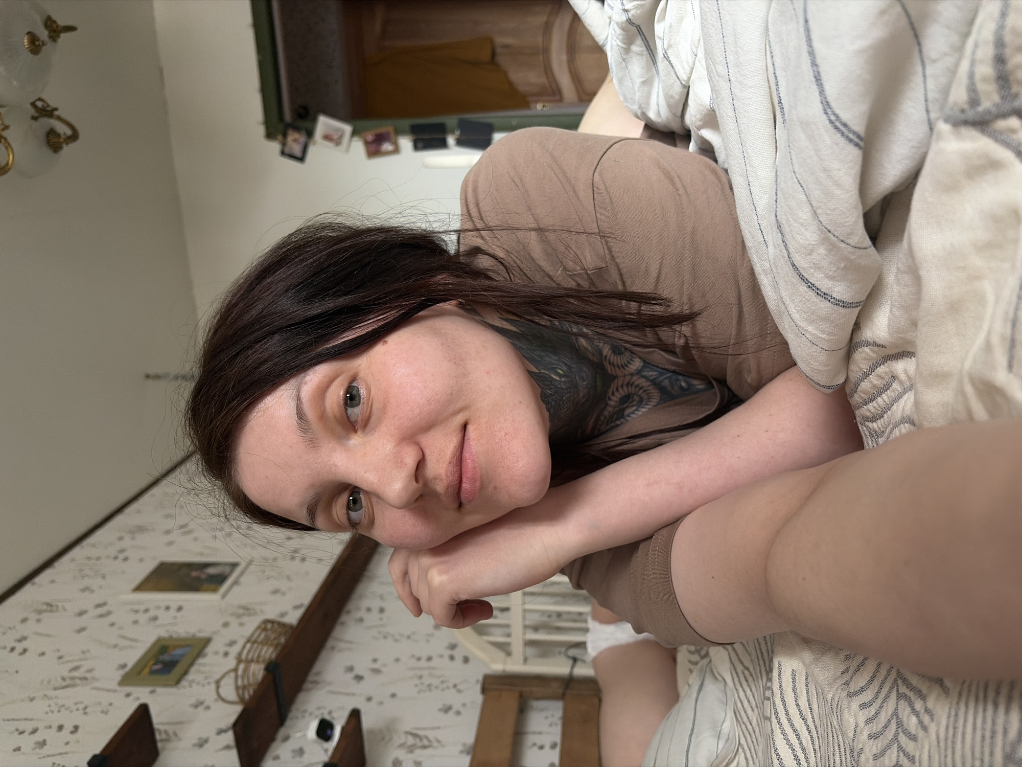 A female, appearing to be between 20 and 30 years old, with dark brown hair and light-colored eyes, lies prone on a bed. She is wearing a plain light brown short-sleeved t-shirt and has an intricate dark-ink tattoo visible on her neck and upper chest. Her head is propped on her clasped hands, and she looks directly at the camera with a neutral expression.
The scene is an indoor bedroom located in the city of Chaikovsky, Russia. The wall behind her on the left is covered with light-colored wallpaper featuring a subtle botanical pattern. Two dark wooden floating shelves are mounted on this wall; the upper shelf holds a small white electronic device, and the lower shelf contains a woven basket. Two framed pictures are also visible on this wall. To the right, a dark green-framed doorway leads to another room, where several small framed photos are affixed to the wall and a yellow garment hangs. A portion of a decorative ceiling light fixture with white globe shades and gold embellishments is visible at the top.
The bed is covered with off-white bedding featuring thin, dark blue horizontal stripes. A wooden headboard is partially visible behind the subject. To her left, behind her arm, a white crib or baby bed is partially visible, containing an infant wearing a white diaper, with one leg exposed.