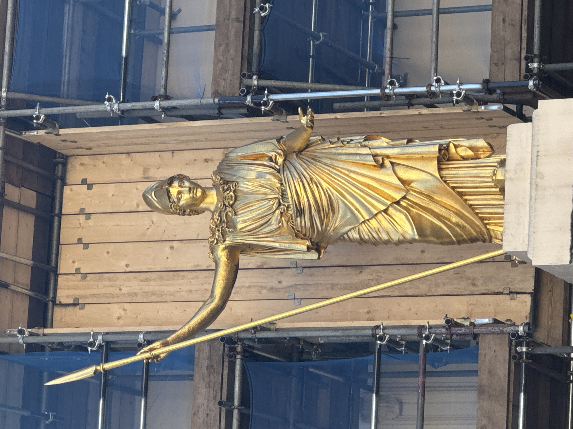 The image shows a golden statue of a woman resembling Athena, the Greek goddess of wisdom, handicraft, and warfare. The statue is being repaired or maintained and is surrounded by scaffolding covered with blue netting. The statue is holding a spear in one hand and raising the other. It appears to be placed on wooden boards which are part of the scaffolding system. The location is likely in the city of London, United Kingdom, as the statue is on top of the Royal Exchange building in the heart of London.