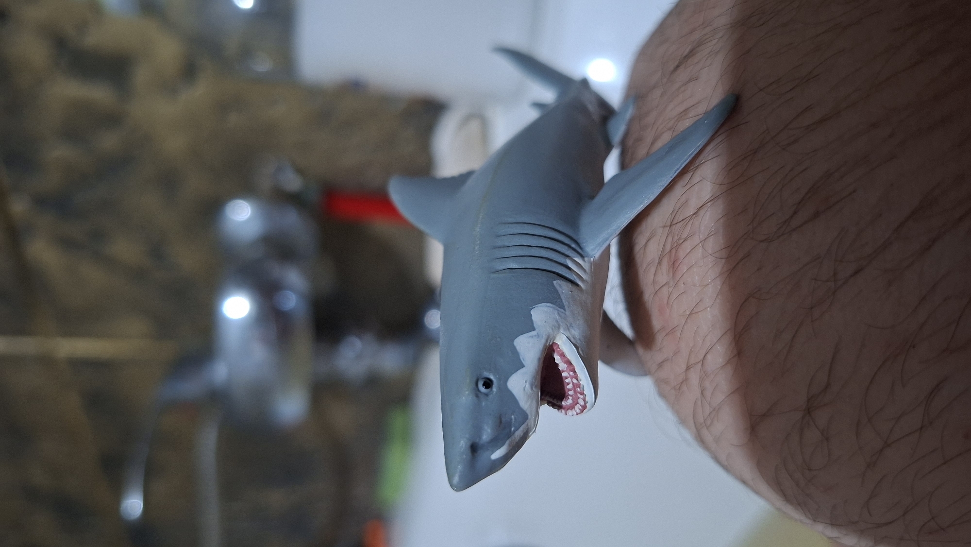 A gray toy shark with an open mouth showing white teeth and pink gums is positioned on a hairy human leg. The shark is on the left side of the frame, facing left. In the blurred background, a faucet with bright reflections is visible, suggesting a bathroom or sink area. The environment appears to be indoors. The location is Voinova, Moldova.