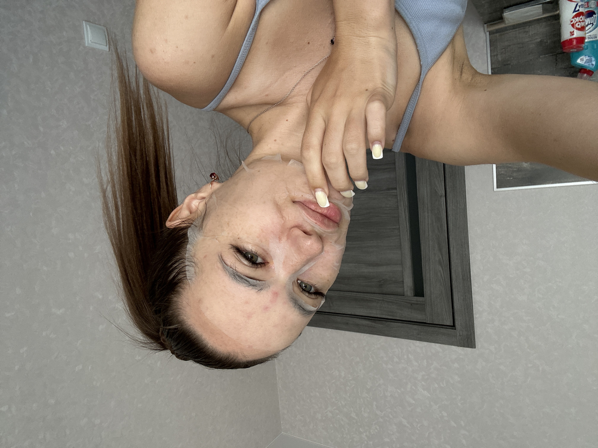 A young woman with a sheet mask on her face and a high ponytail is looking at the camera. She is wearing a light blue tank top and a silver necklace. Her right hand is touching her face, and her fingernails are painted light yellow. She is wearing a small red earring in her right ear. The background is a plain gray wall with a door on the left. The image appears to be taken indoors, possibly in a bedroom.